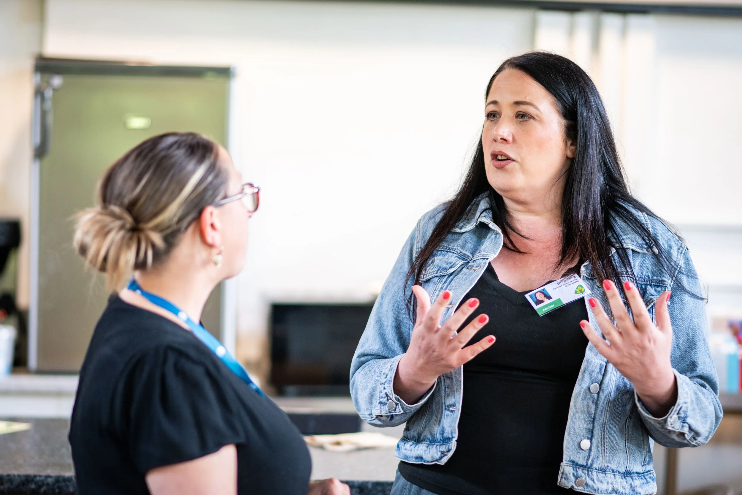 Two women are having a conversation indoors; the woman on the right is talking animatedly, with her hands raised, wearing a denim jacket and a badge, while the woman on the left listens, wearing glasses and a black top.