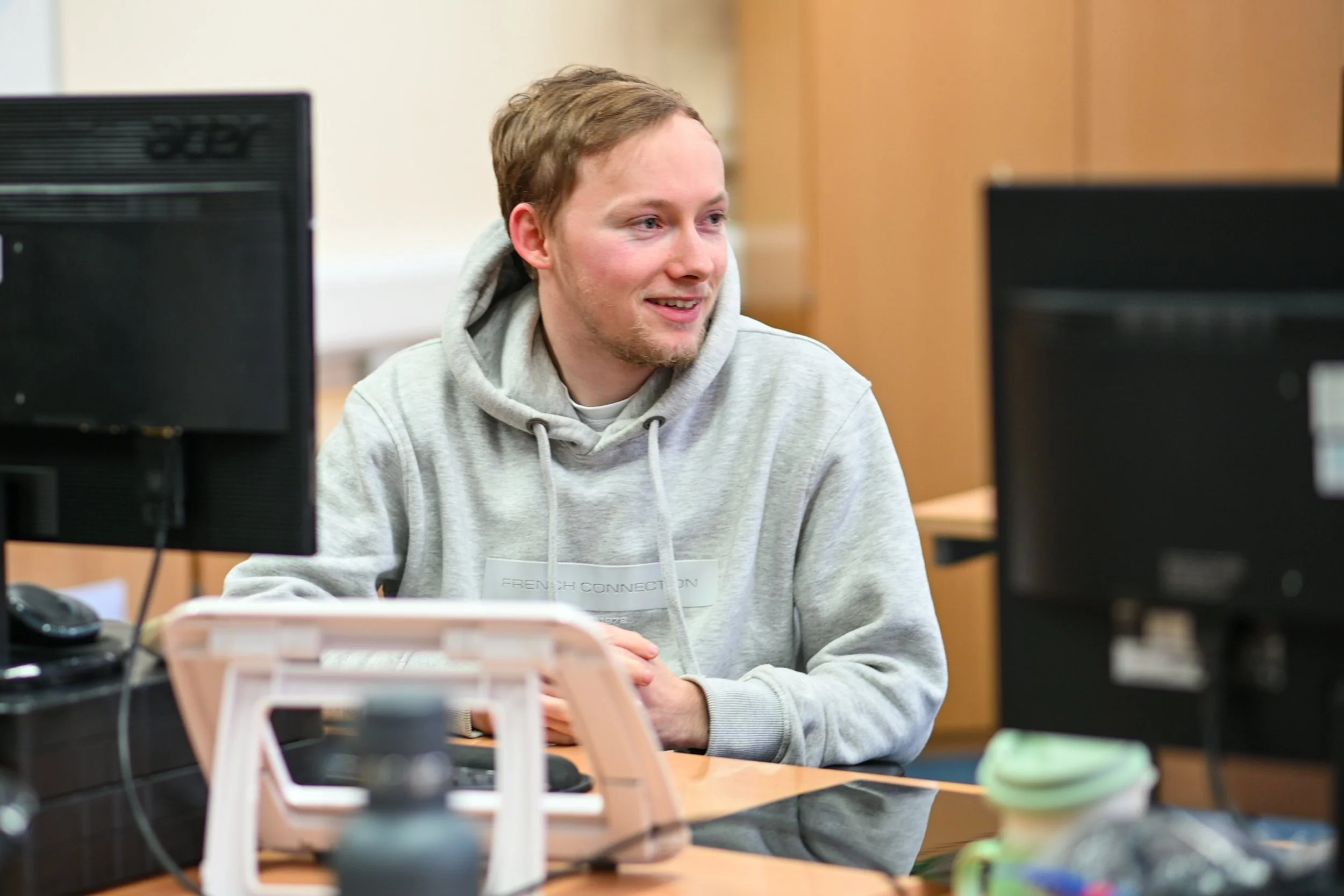 A young man in a gray hoodie smiling and sitting at a desk with two computer monitors in a classroom or computer lab setting.