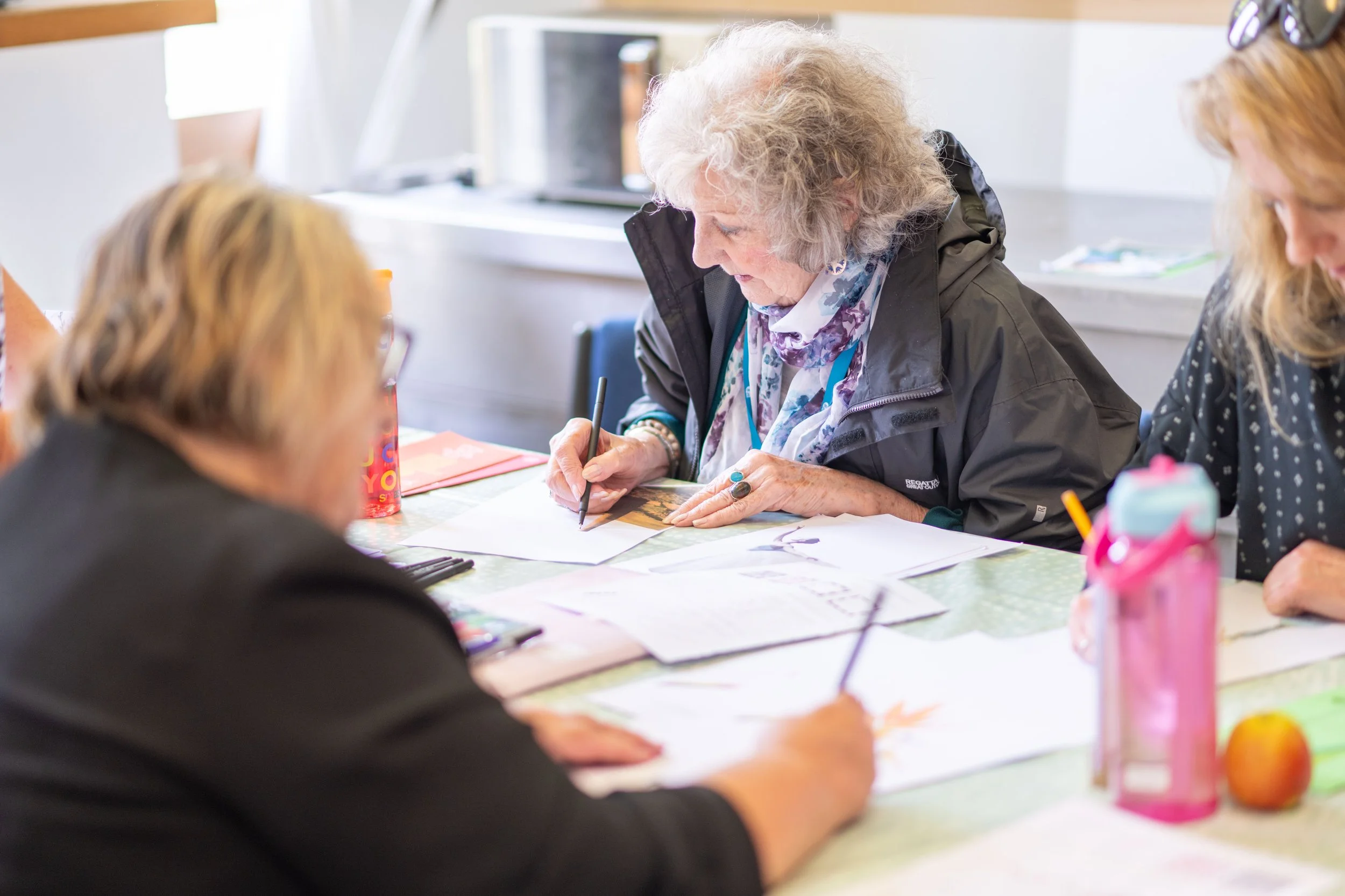 An elderly woman with curly gray hair drawing at a table surrounded by other women, with art supplies and papers on the table.