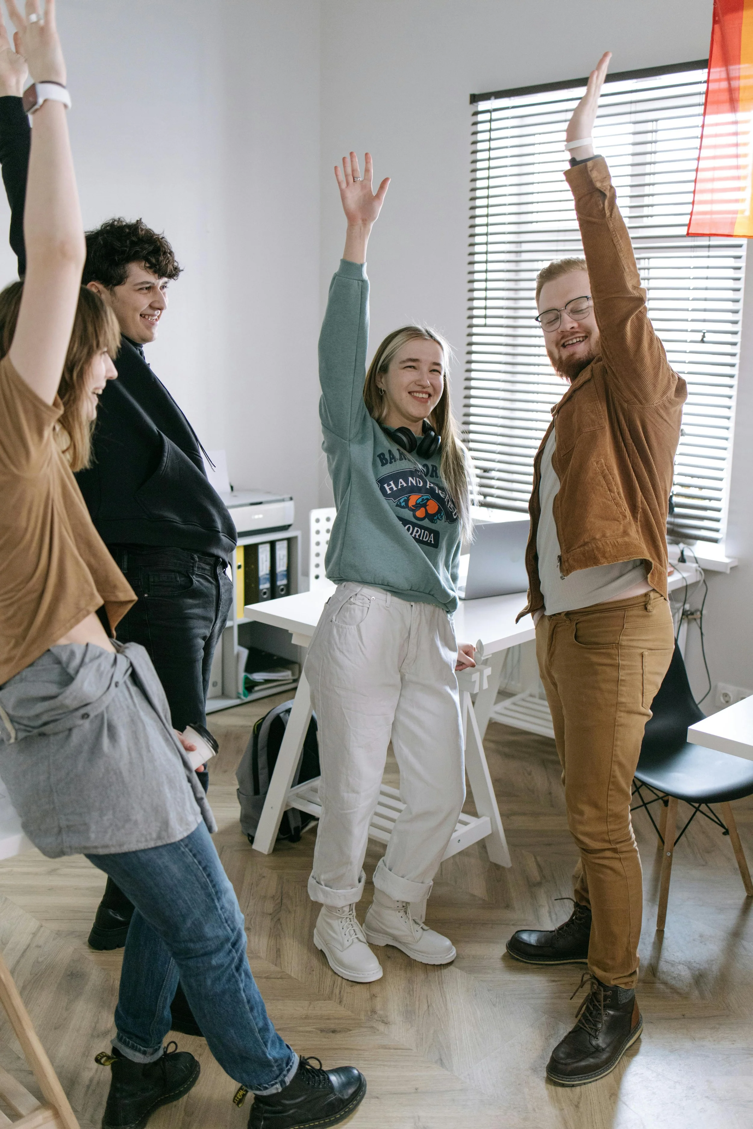 Group of five young adults smiling and raising their hands in a modern office space with natural light from a window.