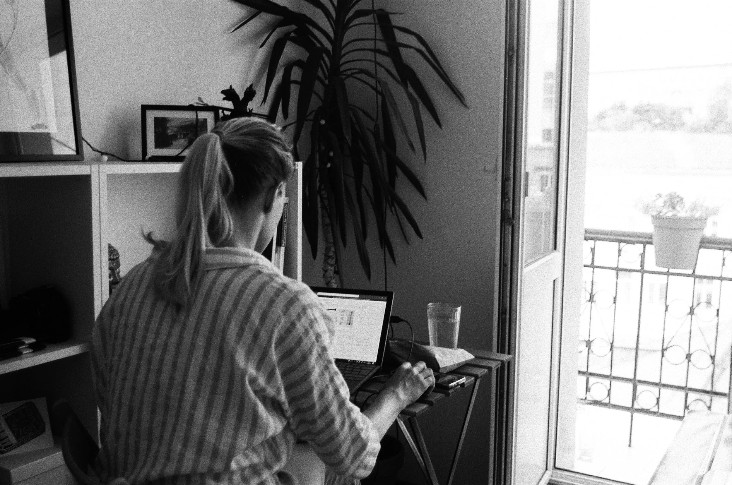 A woman with a ponytail working on a laptop at a small desk near a balcony door, with a glass of water and a phone nearby, in a cozy room with bookshelves and large houseplants.