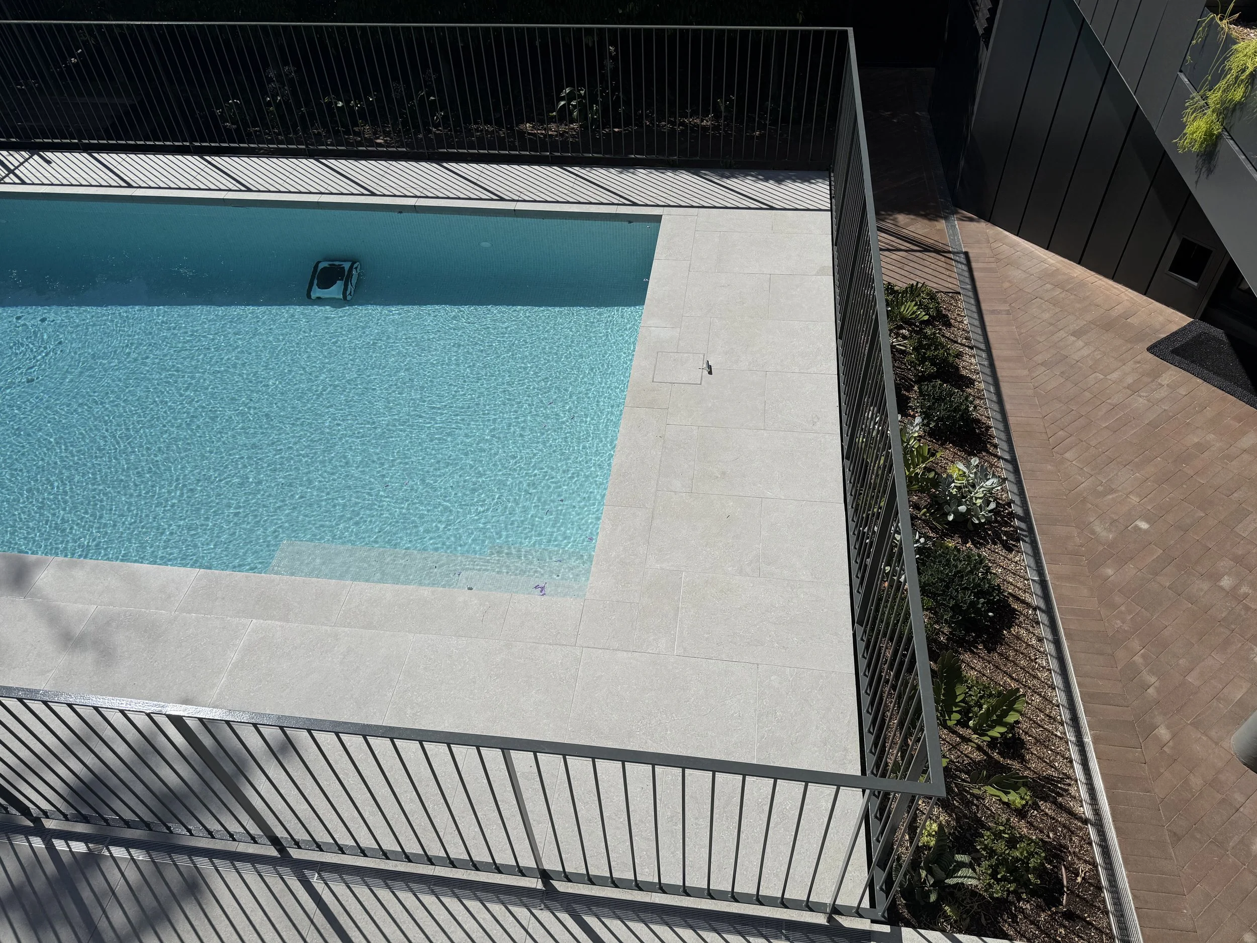 Empty swimming pool with light blue water, surrounded by a grey tiled deck, metal fence, and a garden bed with plants, next to a black building.