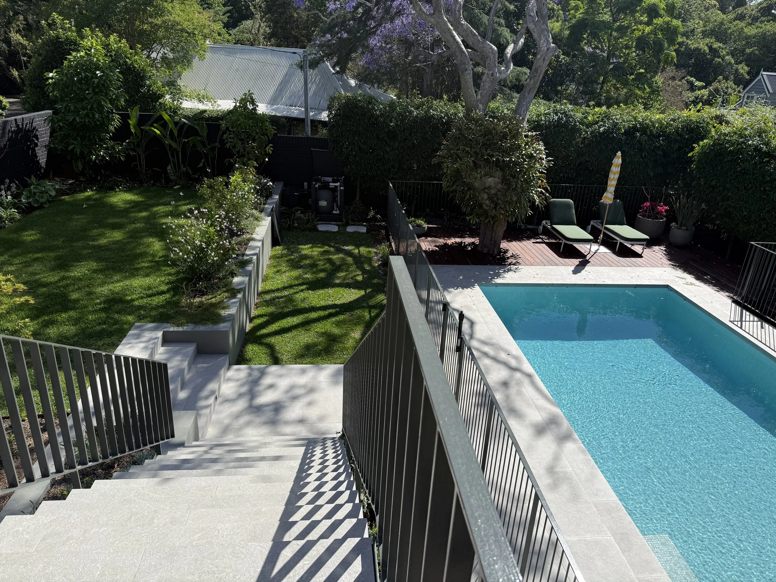A backyard with a swimming pool, green lounge chairs, and a yellow striped umbrella on a wooden deck. There is a large tree and various potted plants. Steps lead down from a patio to the grassy yard surrounded by a black fence.