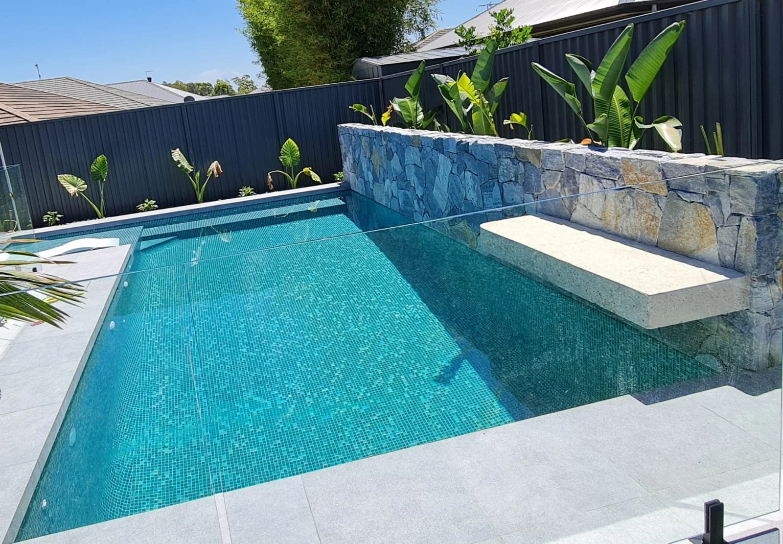 A modern backyard swimming pool with blue mosaic tiles, surrounded by a light gray deck, with a stone wall on one side and a black fence with green plants behind it. Clear blue sky visible overhead.