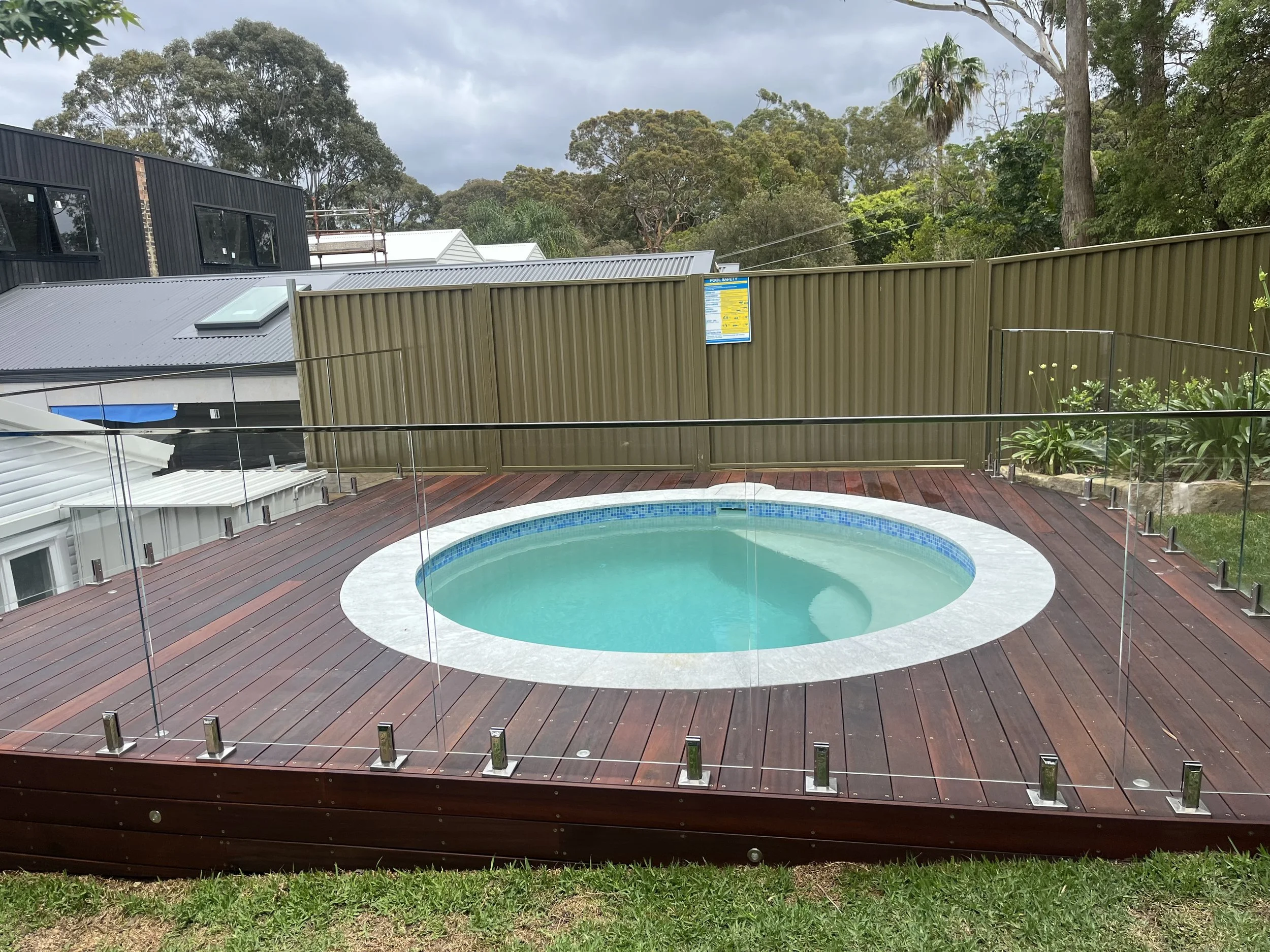 Round outdoor spa with glass fence, wooden deck, and a green backyard with trees and plants.