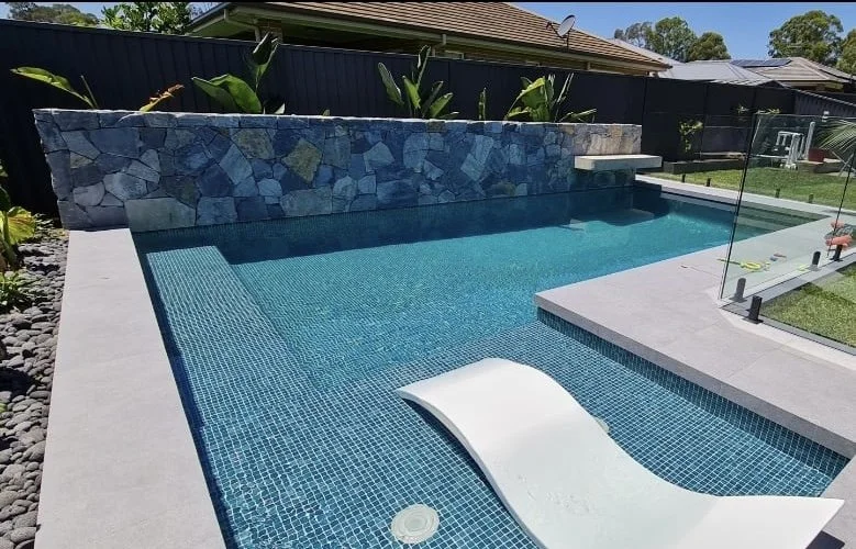 Backyard swimming pool with blue tiles, a small white slide, a stone wall, and a glass fence, with green plants and neighboring houses in the background.