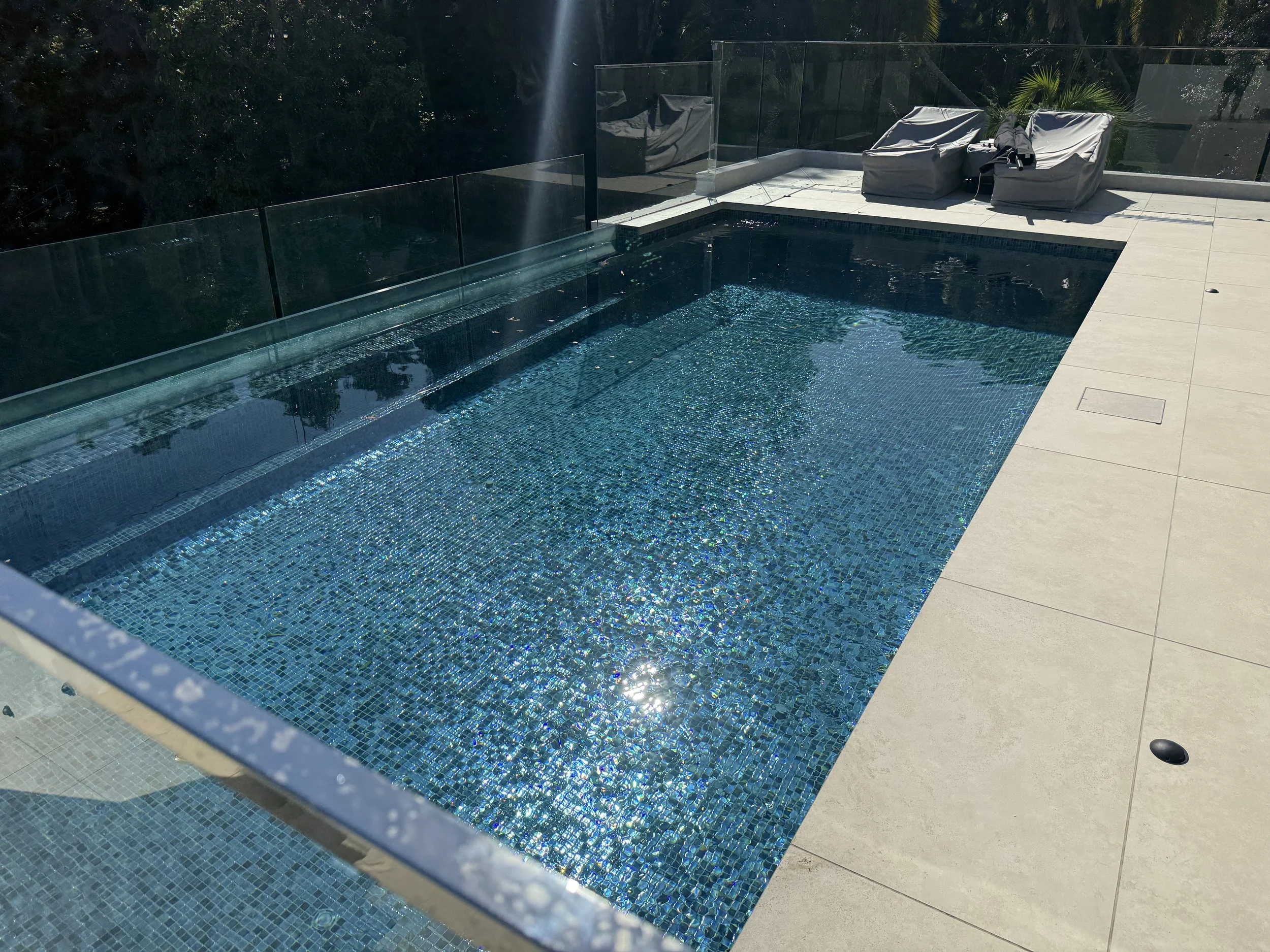 A small rectangular swimming pool with blue mosaic tiles, surrounded by a beige concrete deck. There are two chairs in the background covered with white cloth, and a clear glass fence surrounds the pool area. Sunlight is reflecting on the water.
