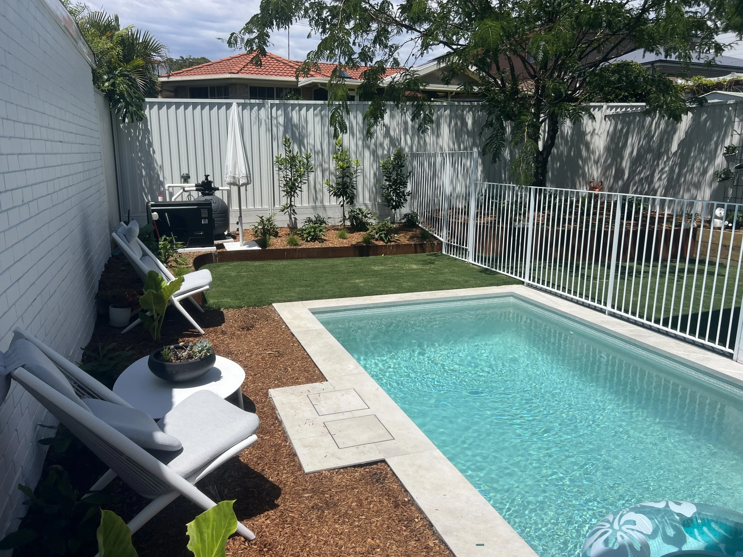 Backyard with a small swimming pool, lawn, patio chairs, potted plants, and a fenced garden with trees and bushes.