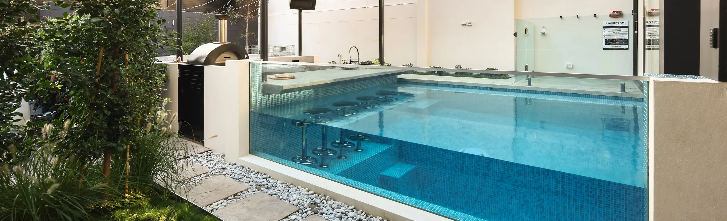 Indoor glass-enclosed pool with built-in seating and hot tub, surrounded by plants and patio stones.
