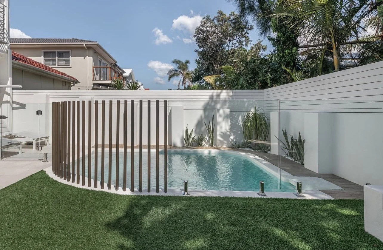 Modern backyard with a small swimming pool, wooden privacy screen, glass fence, green grass, and lush tropical plants against a white wall, under a blue sky with some clouds.