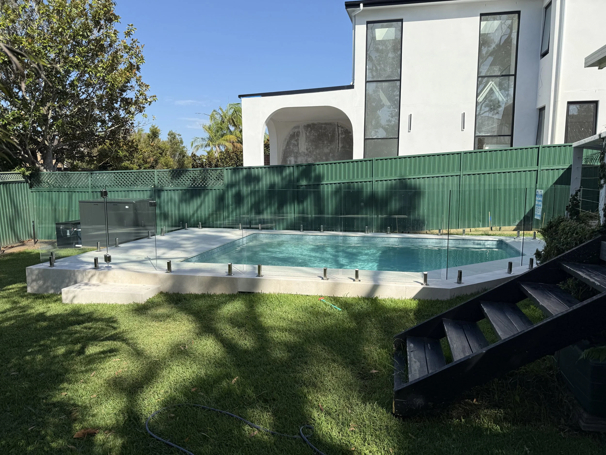 A backyard swimming pool surrounded by a glass fence, with a modern white house in the background and green grass in the foreground.