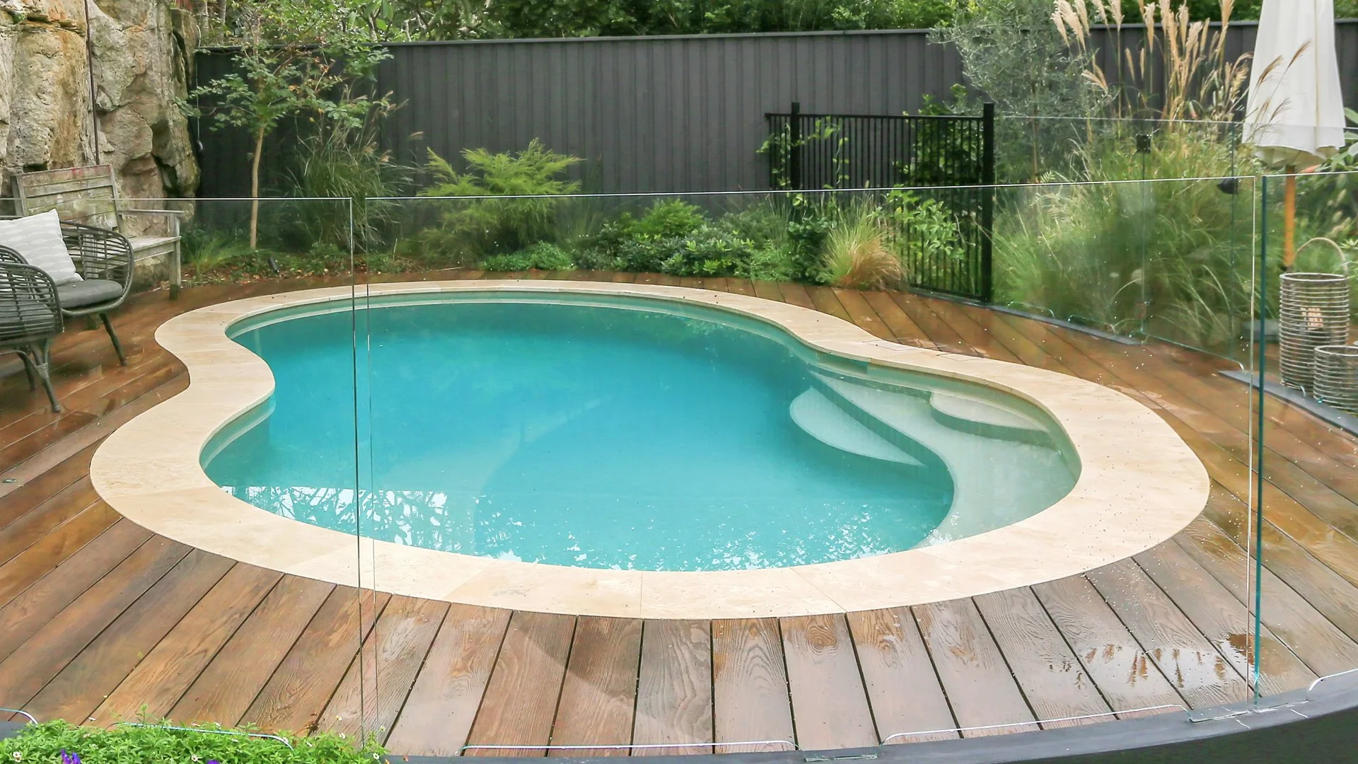 Residential backyard swimming pool, surrounded by a wooden deck, glass safety barrier, garden plants, and outdoor furniture.