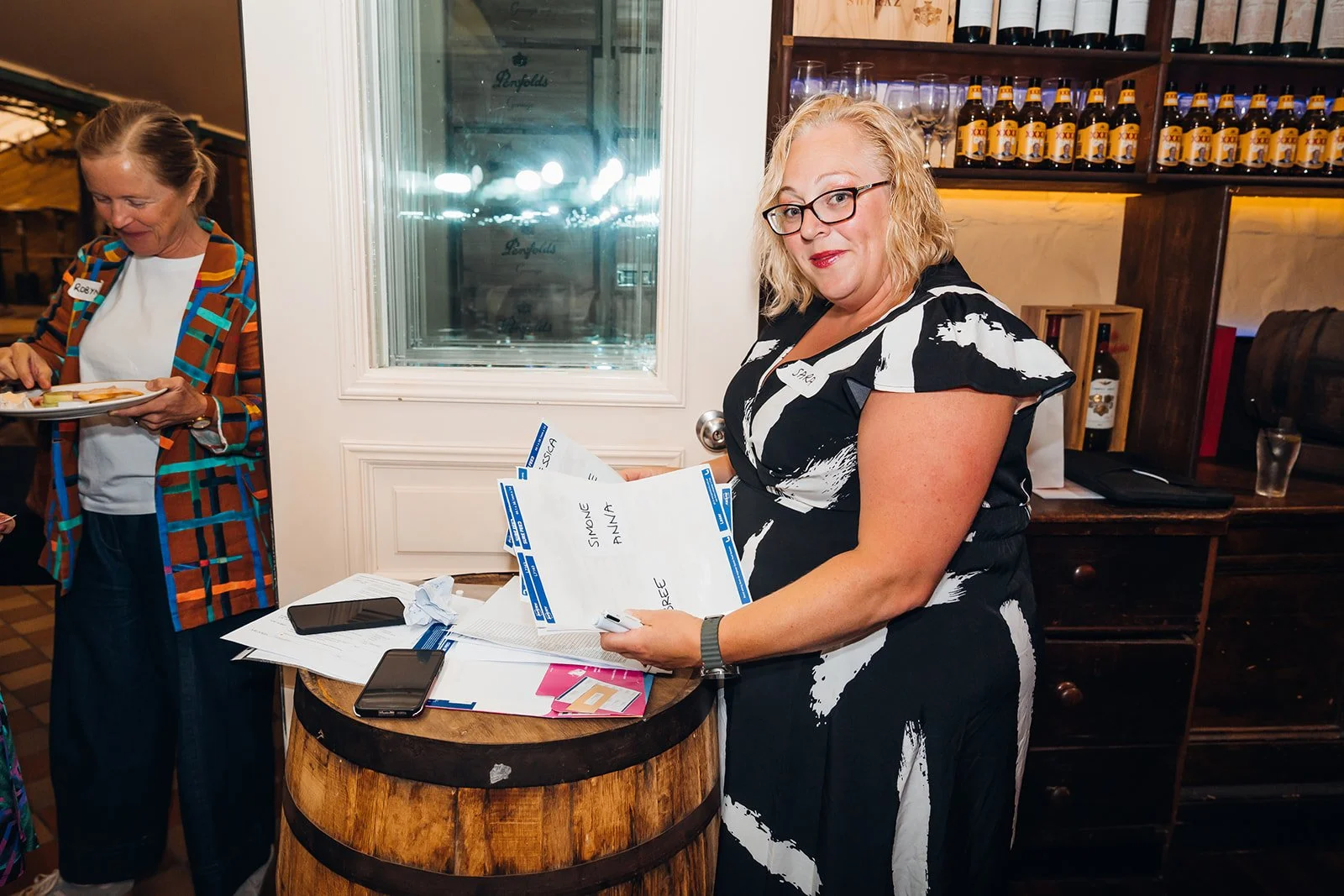 A woman with glasses and a black and white dress holding papers, standing beside a small wooden table with papers and two smartphones on it, in a restaurant or bar setting.