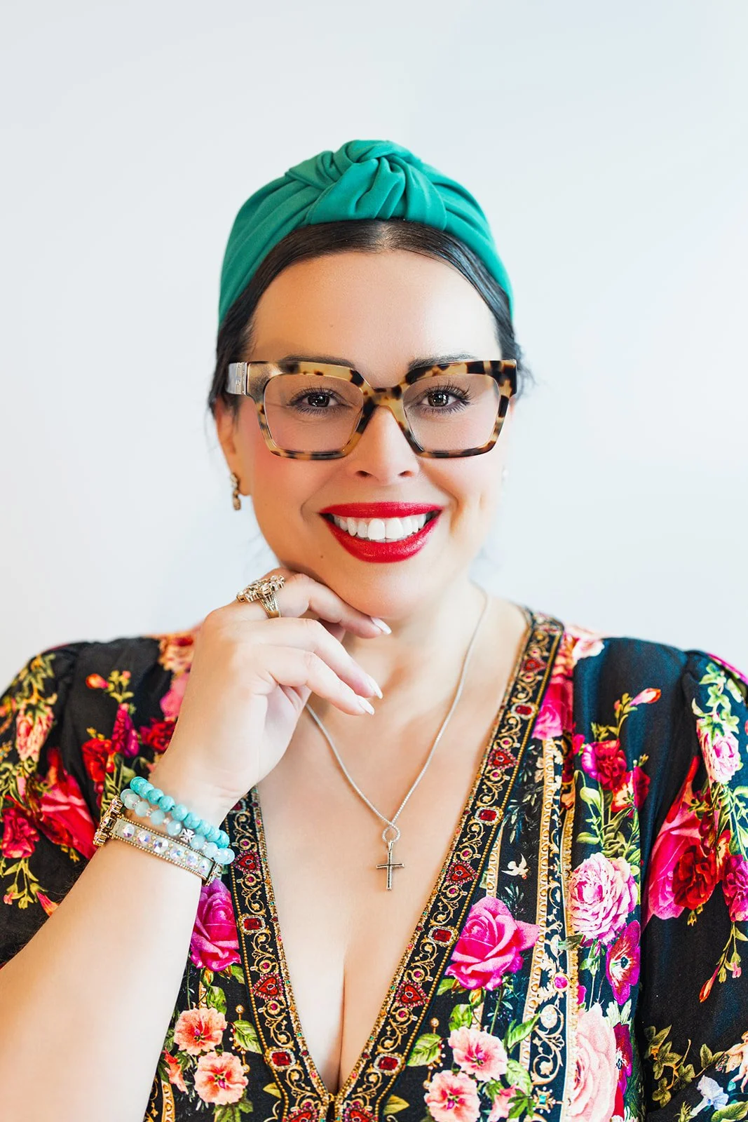A woman wearing glasses with a tortoise pattern, a teal headband, red lipstick, and colorful floral dress, smiling and resting her chin on her hand.