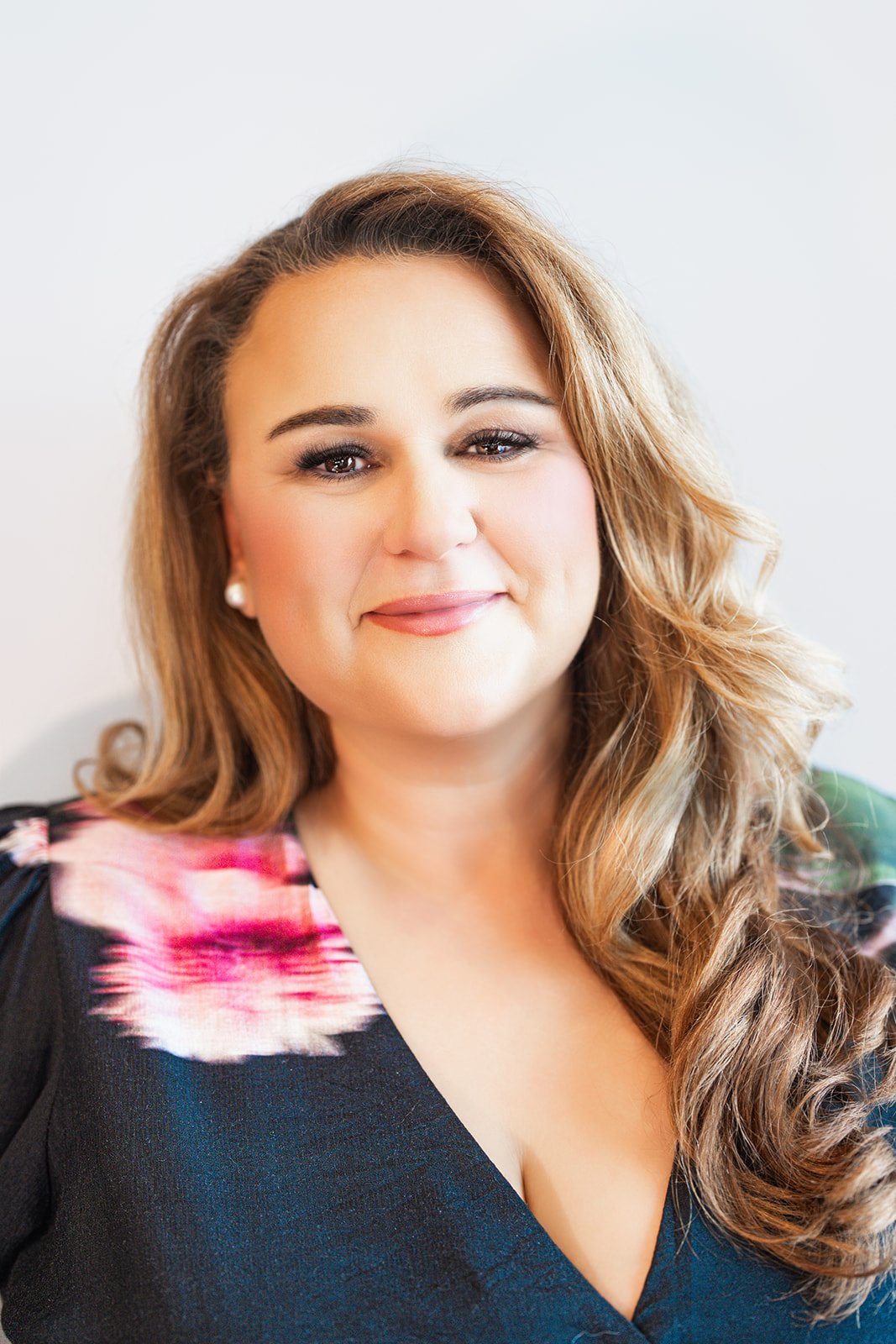 A woman with long wavy hair smiling at the camera, wearing a black top with floral accents on the shoulders, standing against a white background.