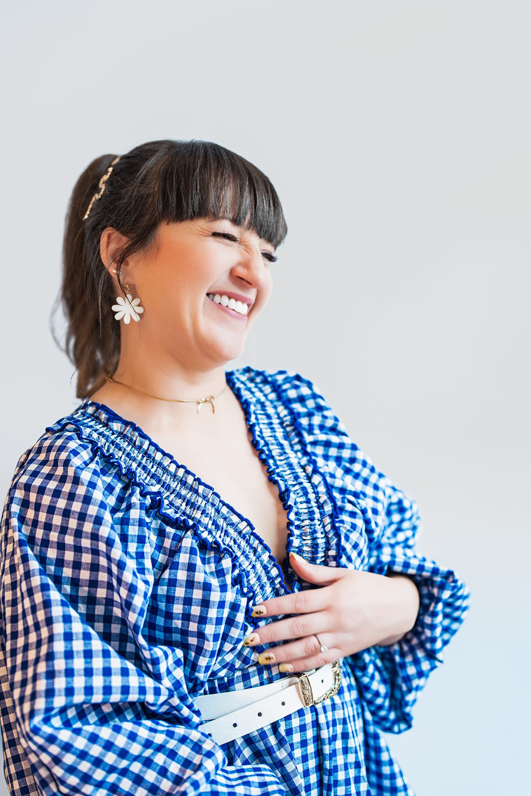 A woman with dark hair in a ponytail, wearing white flower earrings and a blue gingham blouse, smiling and holding her chest with one hand.