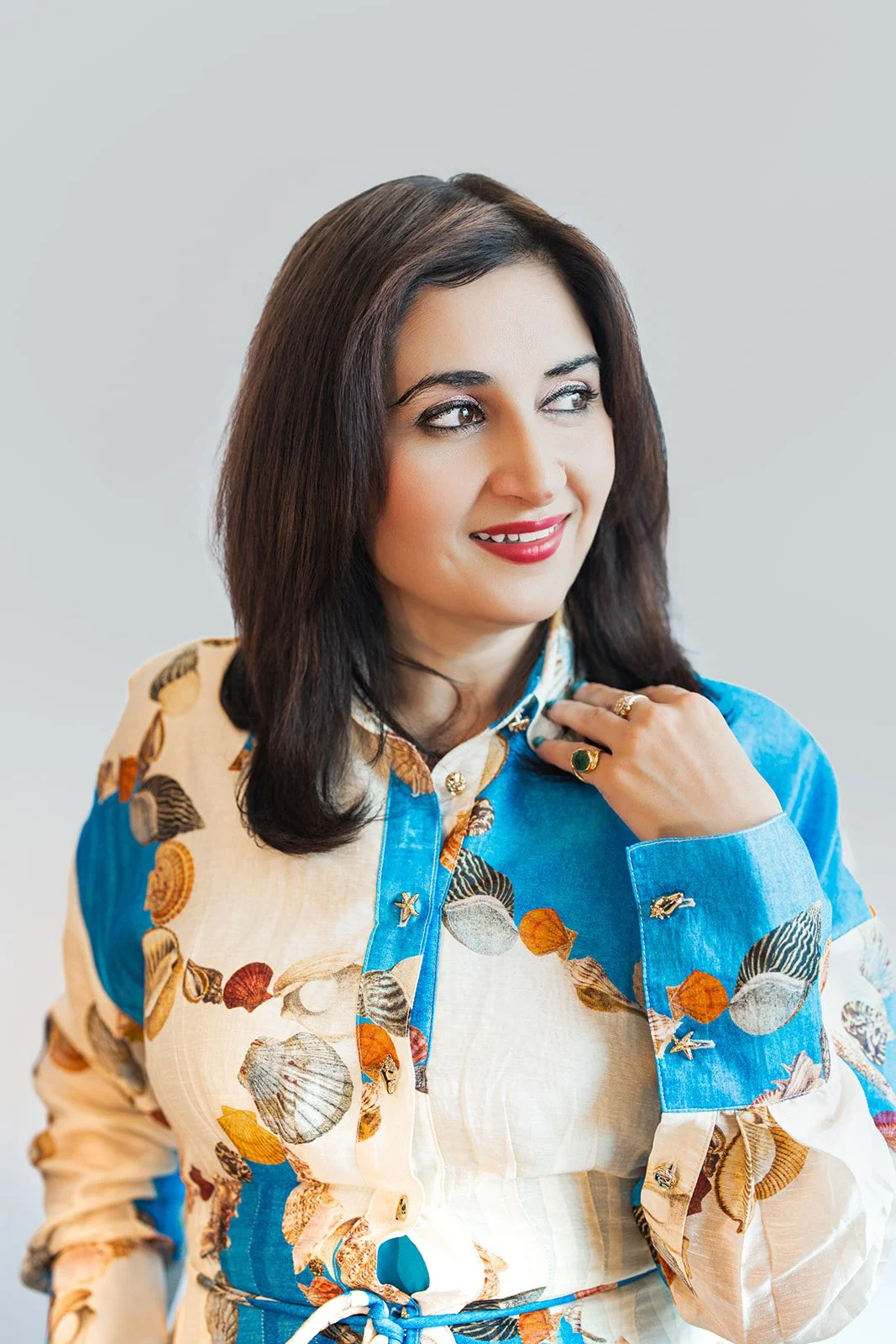 A woman with dark brown hair, wearing a colorful seashell-patterned dress with a blue collar, smiling and looking to her left.