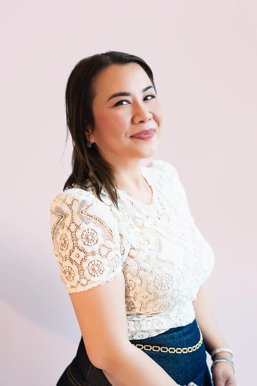 A woman with dark hair styled in loose waves, wearing a white lace blouse with short sleeves and a navy skirt with a chain belt, smiling softly against a plain light pink background.
