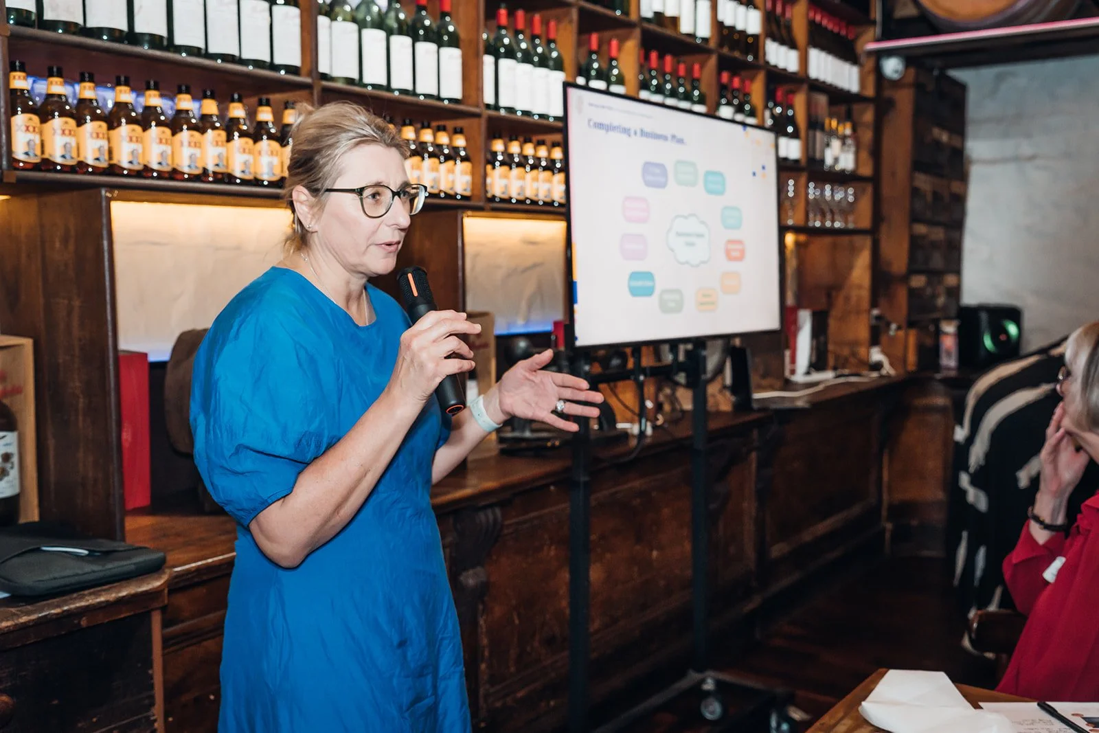 Woman giving a presentation with a microphone in a restaurant or bar setting, people seated at tables, wine bottles on shelves, and a large screen displaying a colorful diagram in the background.
