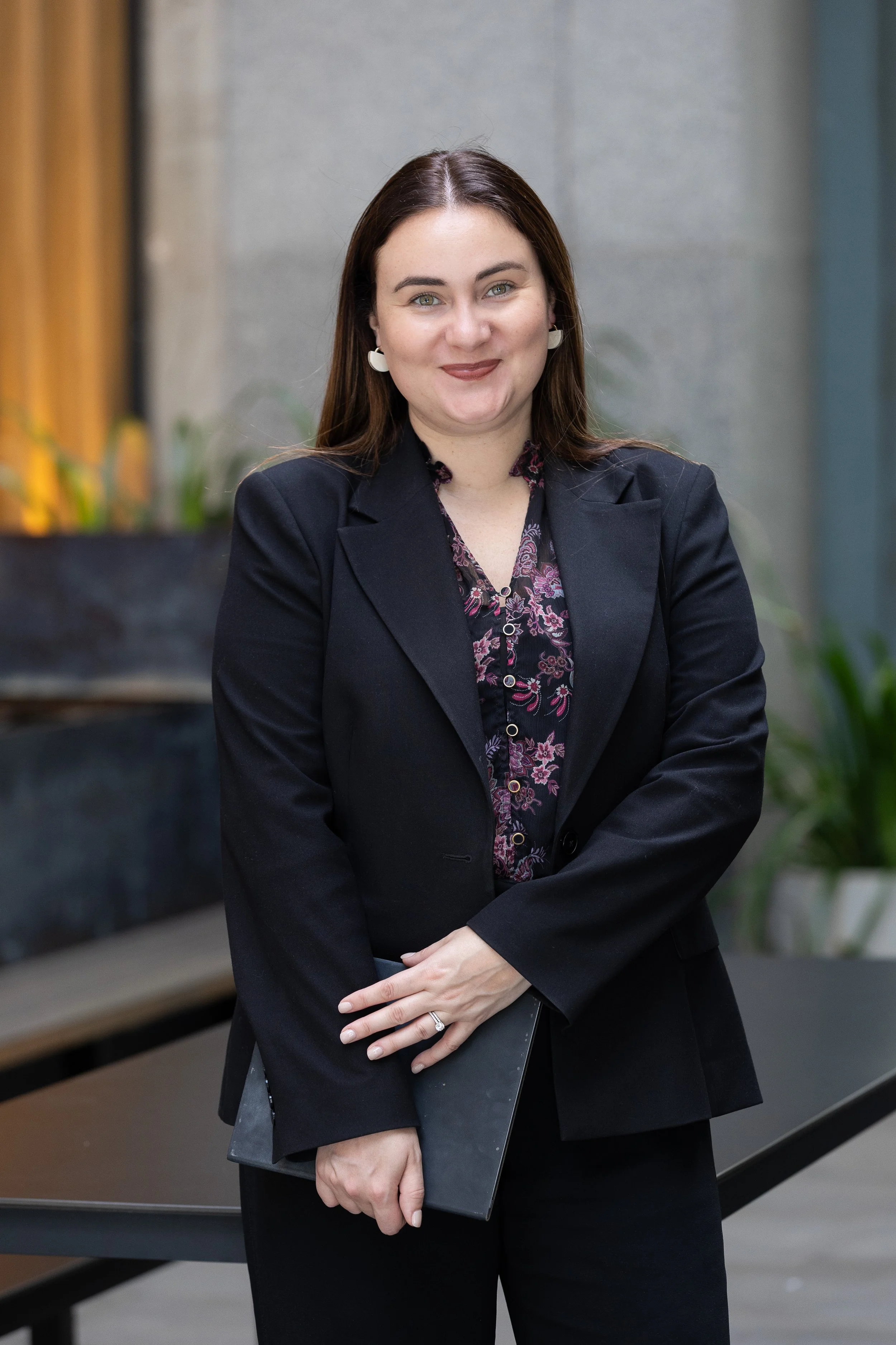 A woman with long brown hair wearing a dark blazer and floral shirt, holding a black portfolio or folder, standing with her arms crossed in an office or professional setting.