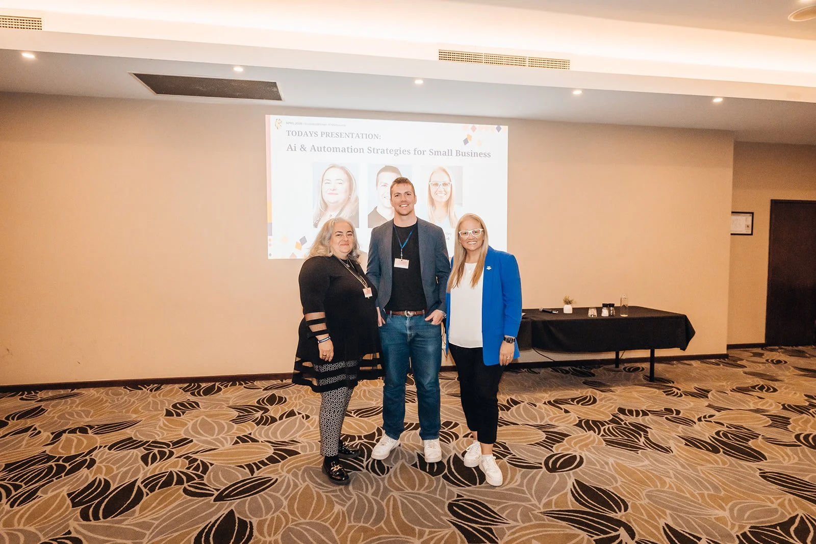 Three individuals standing together in front of a presentation screen at a conference or seminar. The screen displays a presentation titled 'AI & Automation Strategies for Small Business' with images of three people. The setting is a conference room with a patterned carpet and a black table to the right.