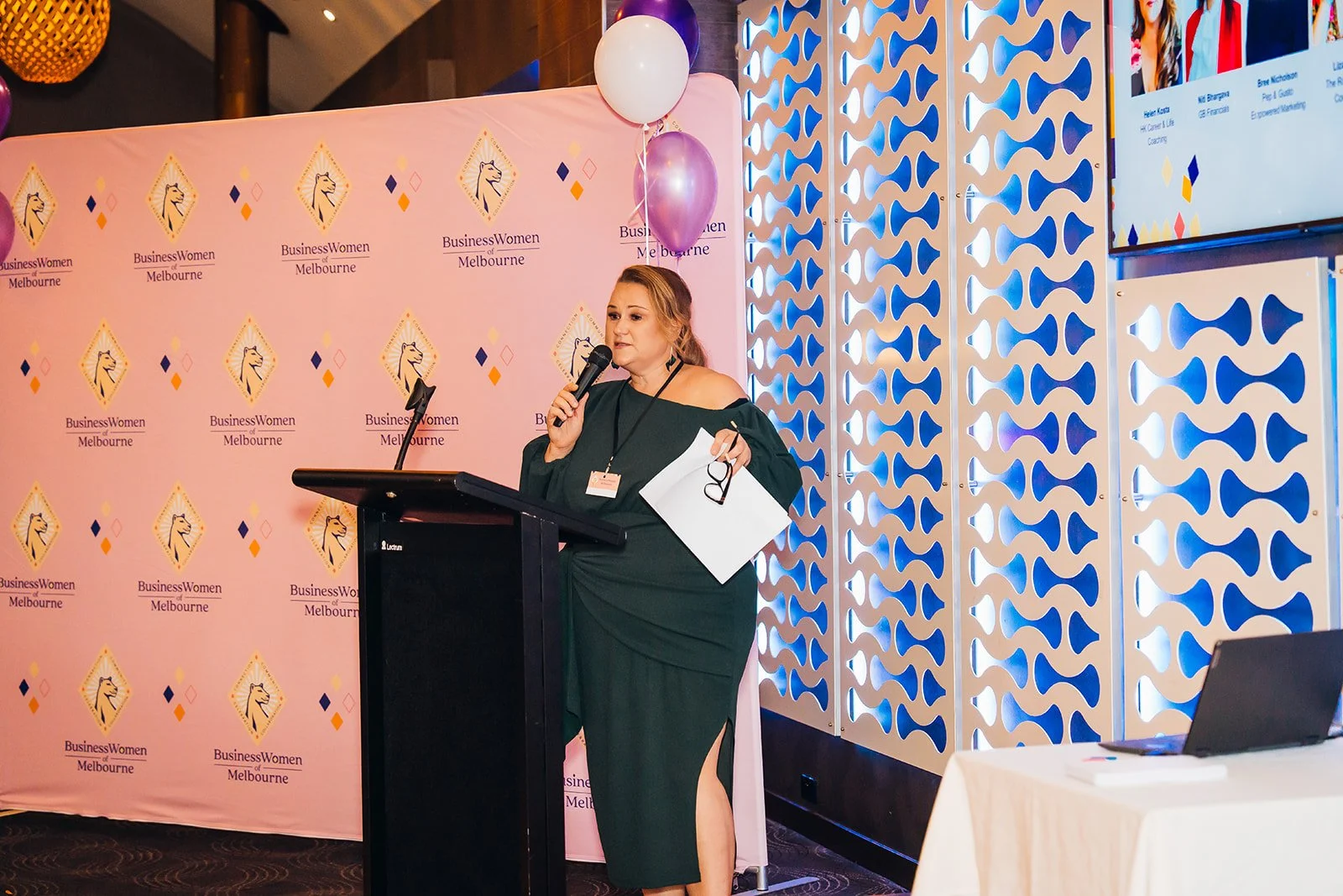 A woman in a dark green dress speaking at a podium with a microphone, holding glasses and papers, backstage at a 'Business Women of Melbourne' event, with balloons and a pink backdrop featuring the event logo.