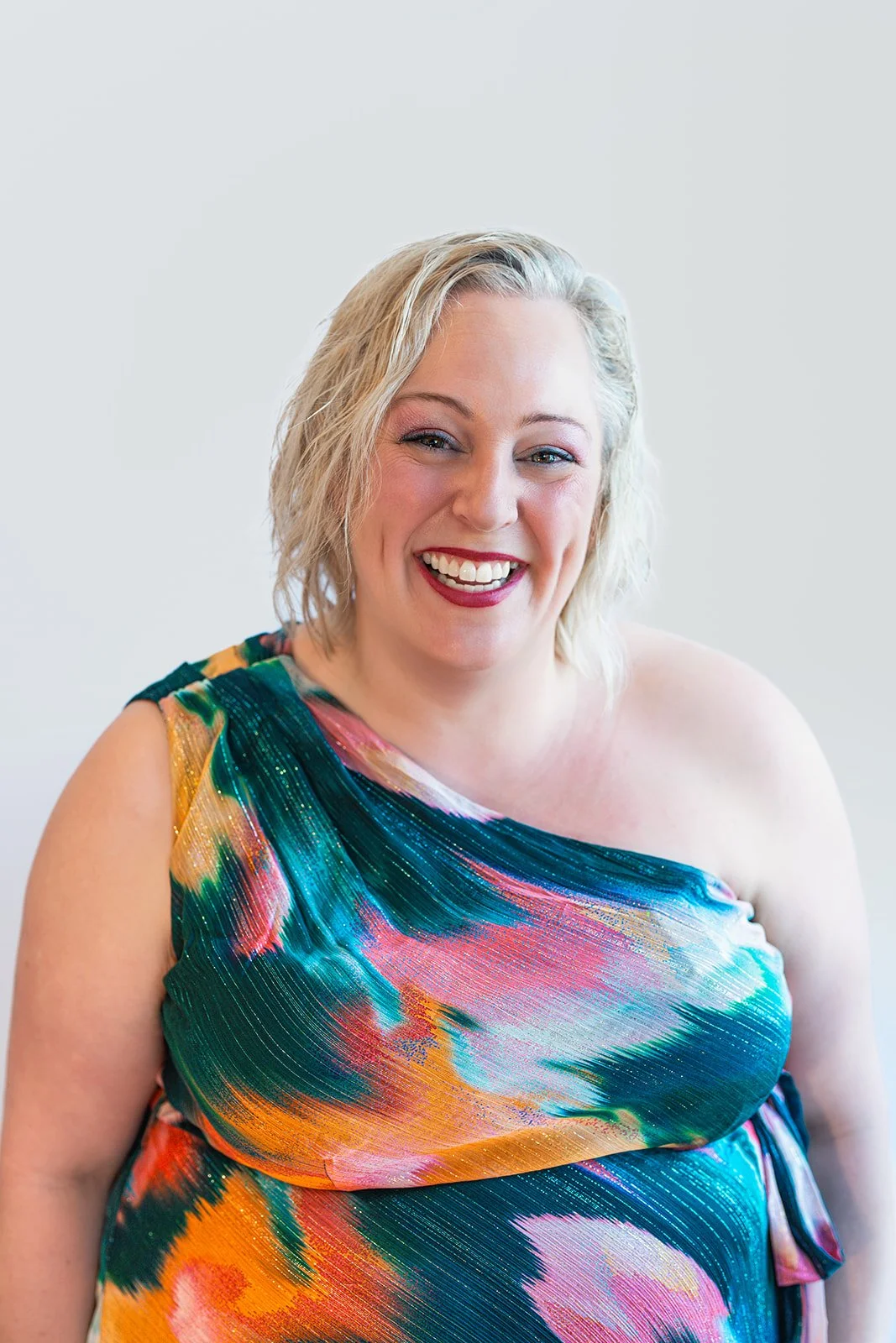 A smiling woman with short blonde hair wearing a colorful, abstract patterned one-shoulder dress.