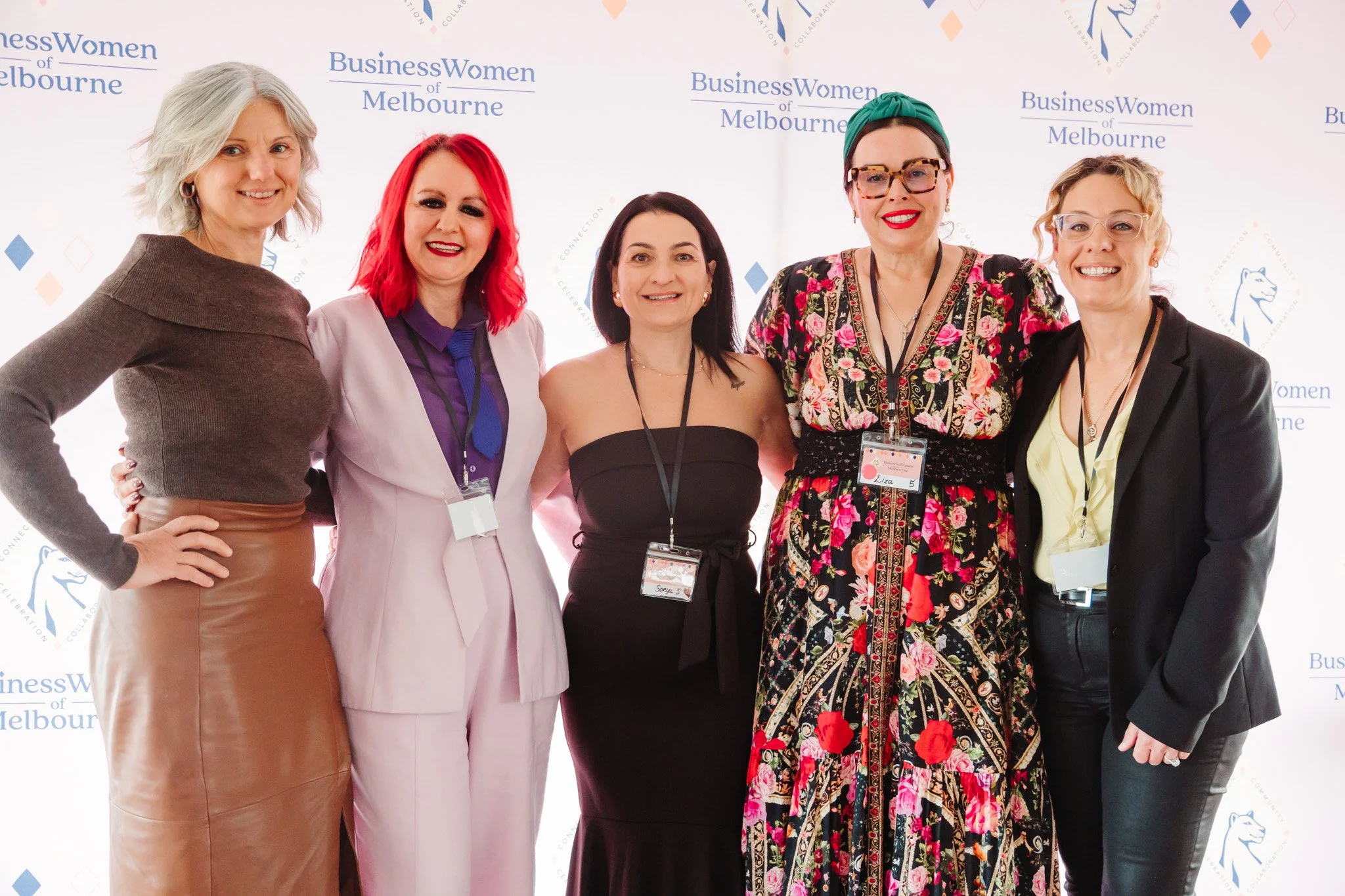 Group of five women standing in front of a backdrop with the text 'Business Women of Melbourne' and a logo. They are dressed in colorful and professional attire, smiling at the camera.