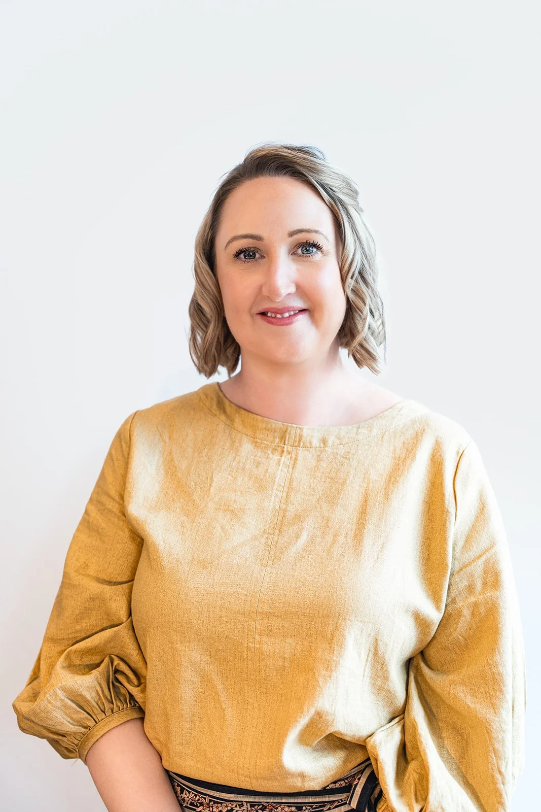 A woman with short, wavy blond hair, smiling, wearing a mustard yellow top with three-quarter length sleeves and a patterned skirt, standing against a plain white background.