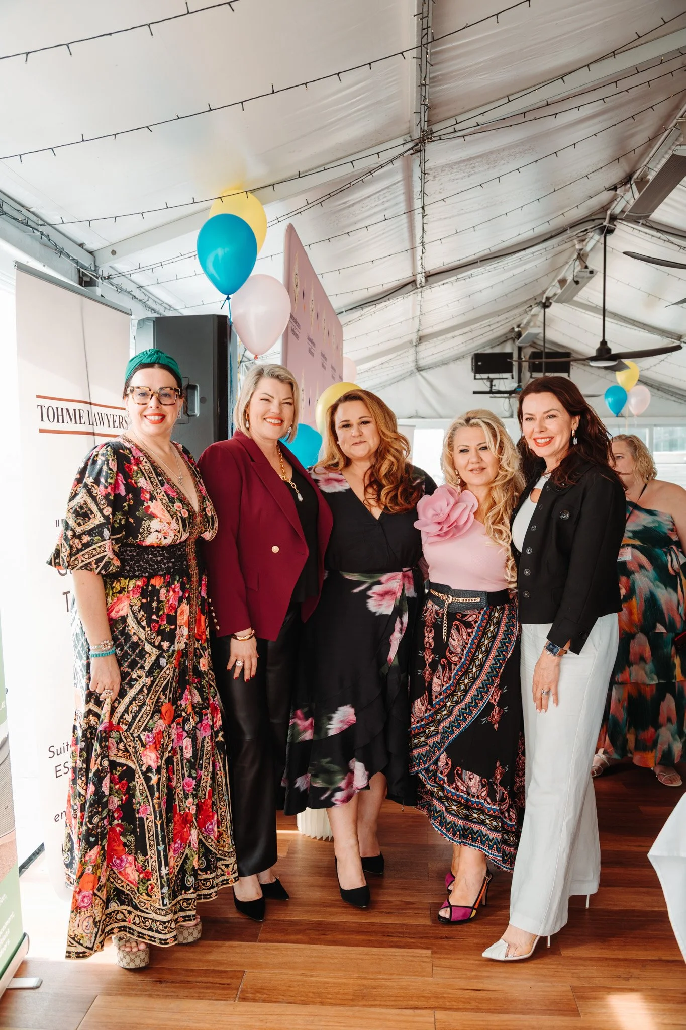 Group of six women standing together inside a large tent decorated with balloons, smiling at the camera, dressed in colorful and stylish clothing.