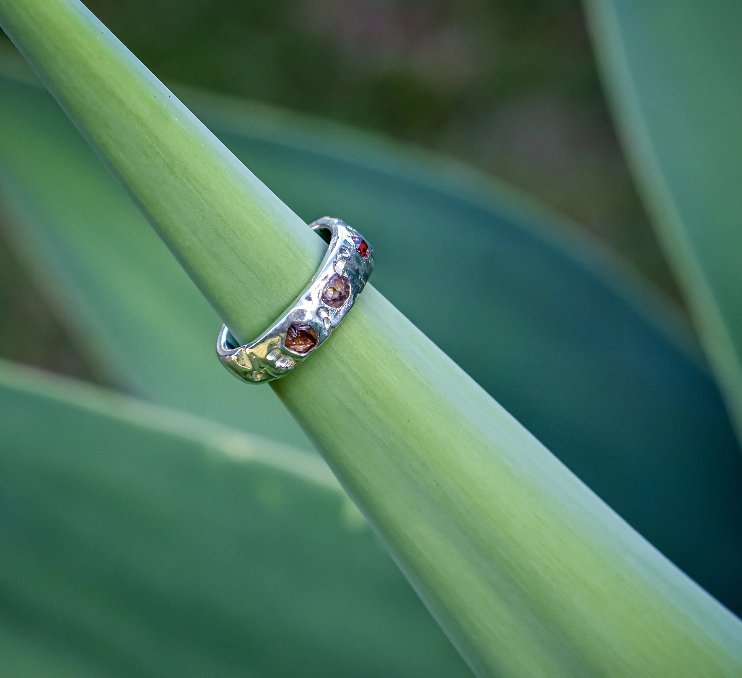 A silver ring with embedded colorful stones resting on a green plant stem.