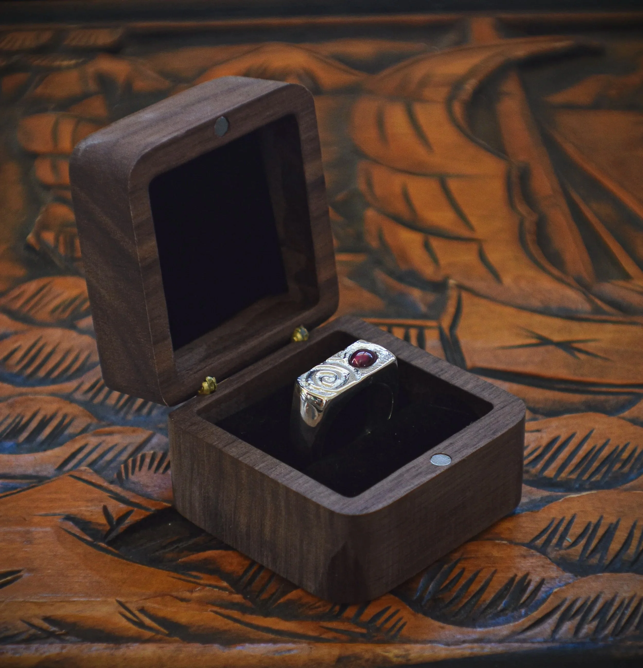 A silver ring with a red gemstone inside a wooden jewelry box, on a carved wooden surface.