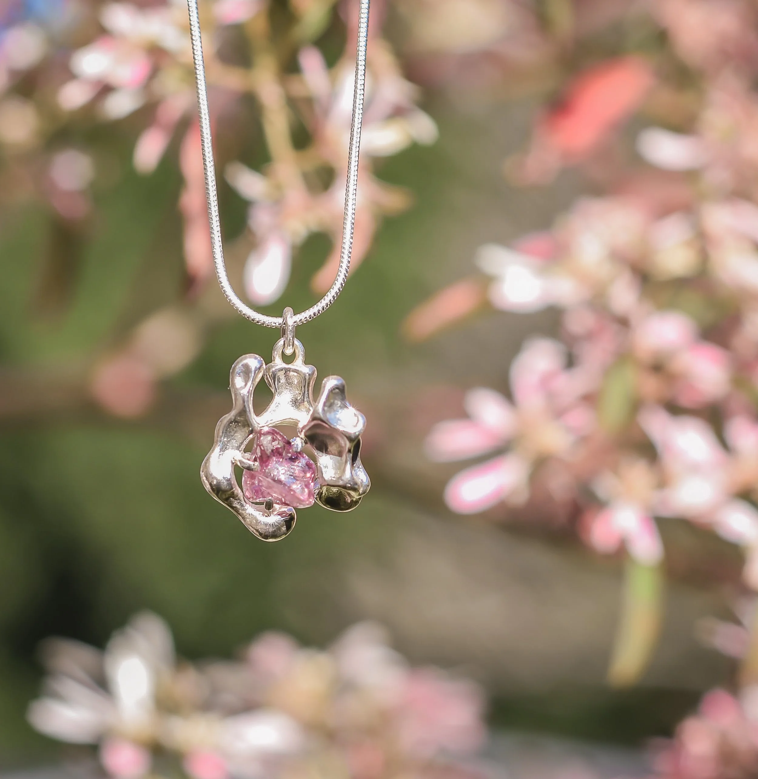 Silver necklace with a pendant featuring an abstract, organic-shaped silver setting with a pink gemstone in the center, hanging in front of a blurred background of pink and white flowers.