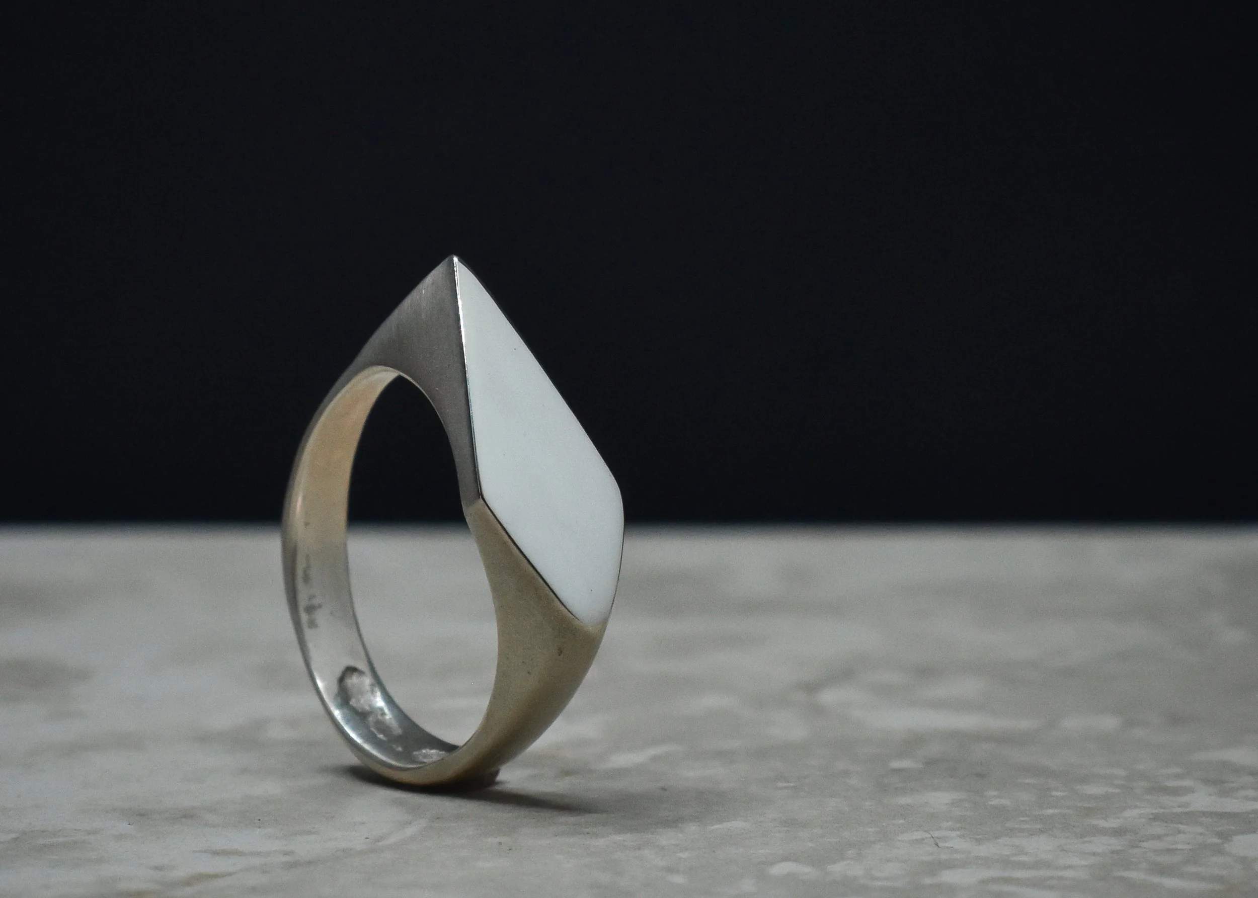Close-up of a silver ring with a large, flat geometric top, placed on a flat surface against a black background.