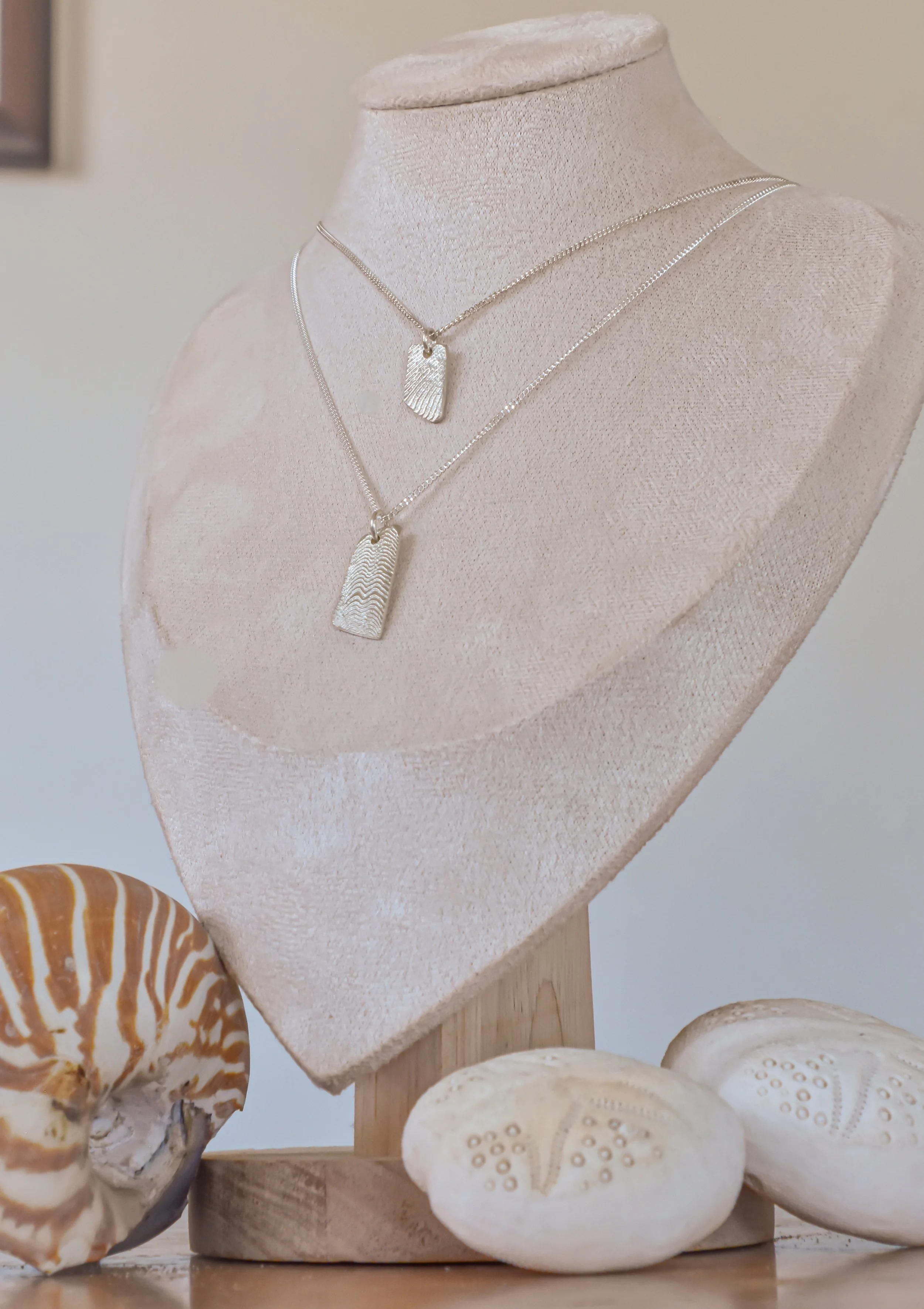 Two layered silver necklaces with rectangular pendants on a mannequin neck, with seashells in front of it.
