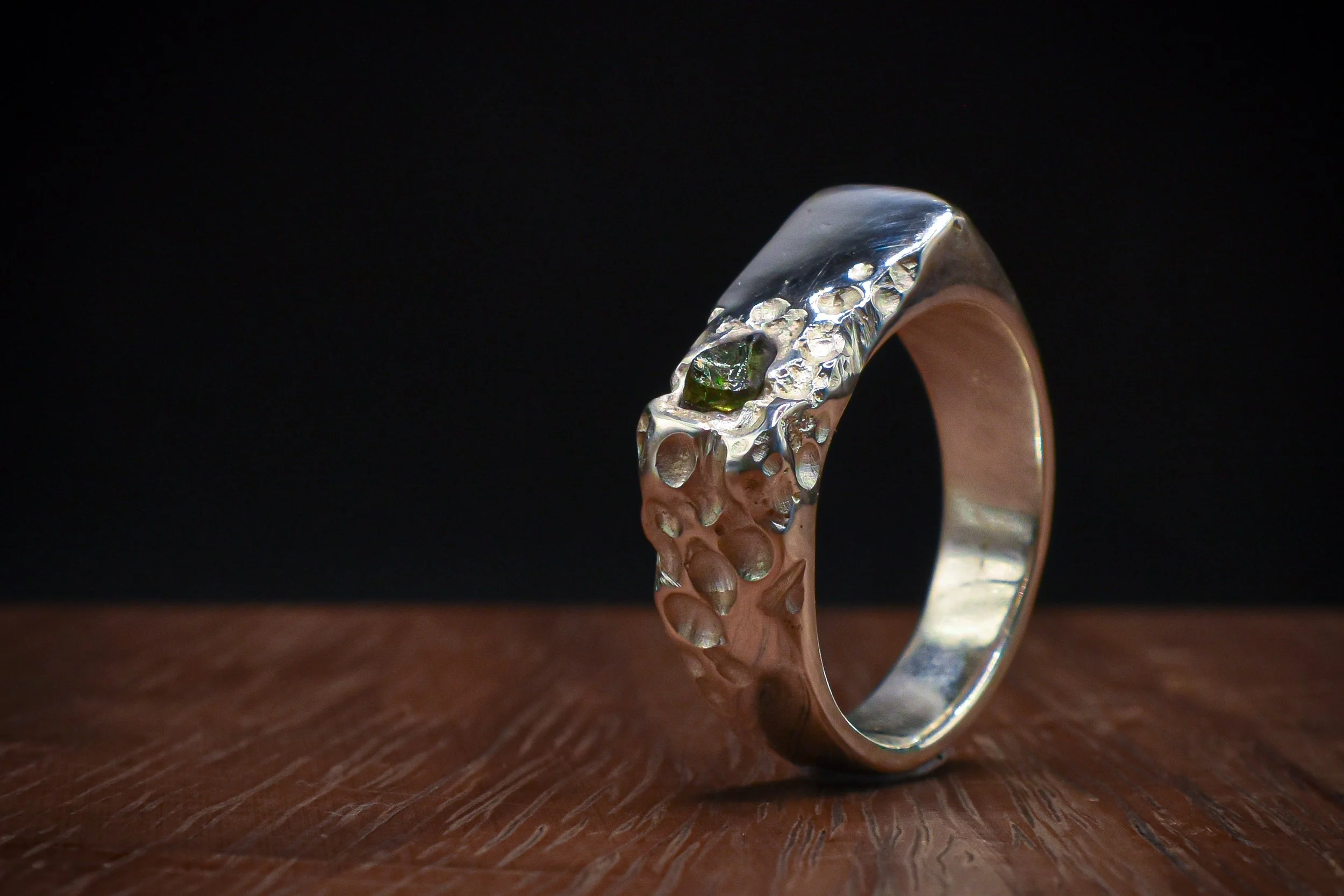 A silver ring with textured surface and small green gemstone on a dark background, placed on a wooden surface.