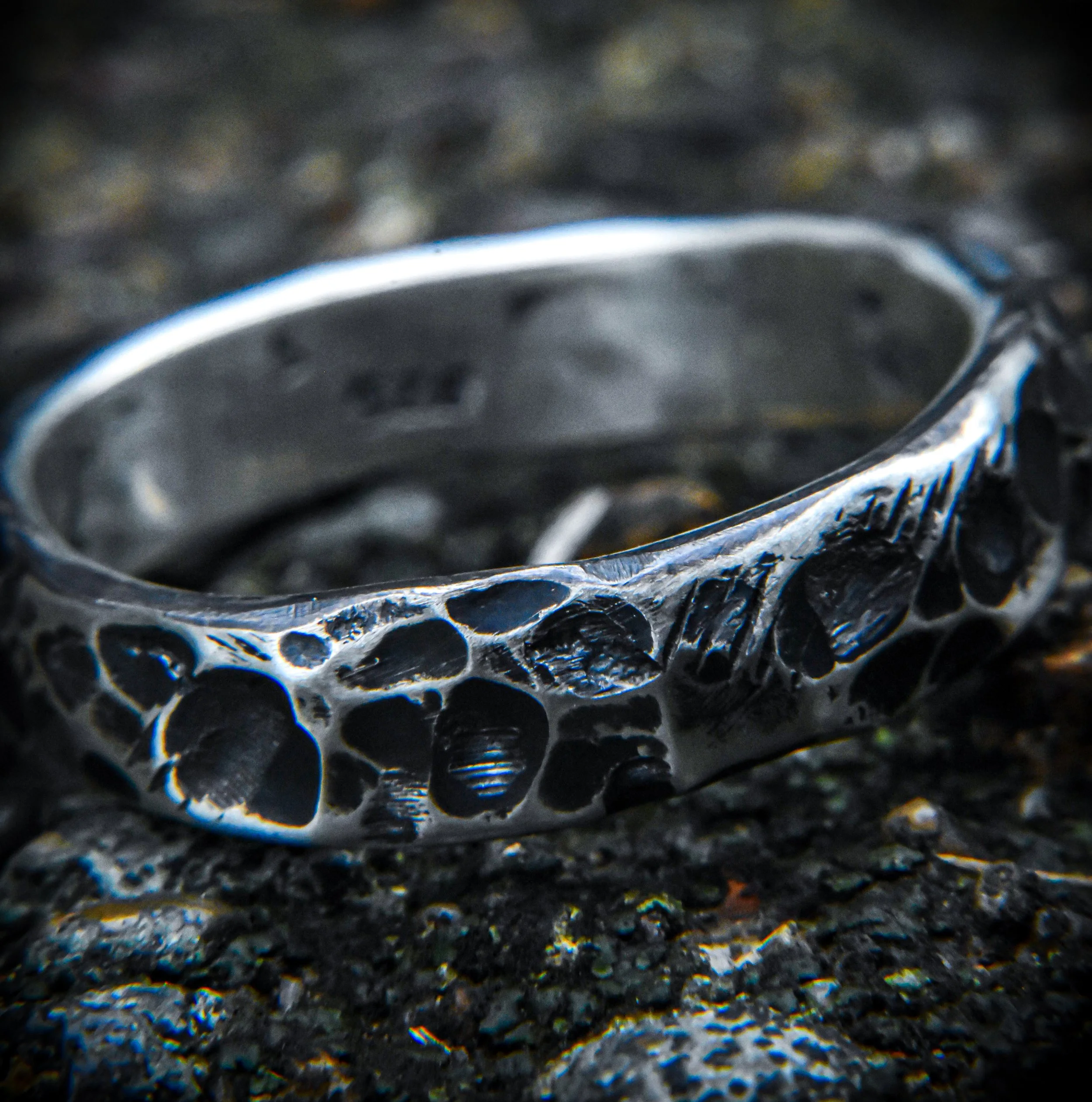 Close-up of a silver ring with a hollow, intricate design resting on dark, textured ground.