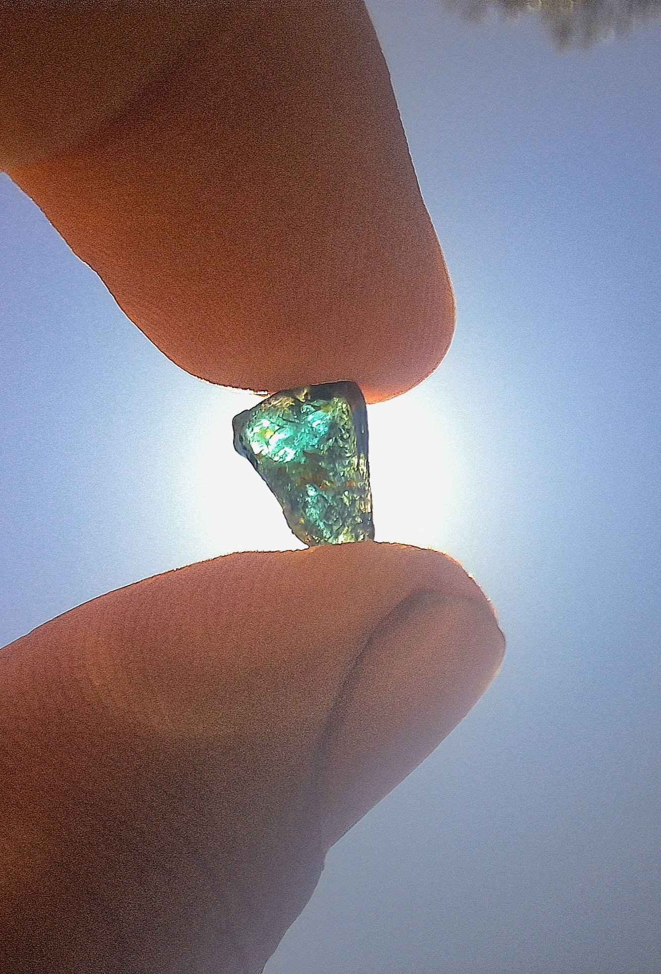 Close-up of a person's fingers holding a small, irregularly shaped opal gemstone against the bright sun in the sky.