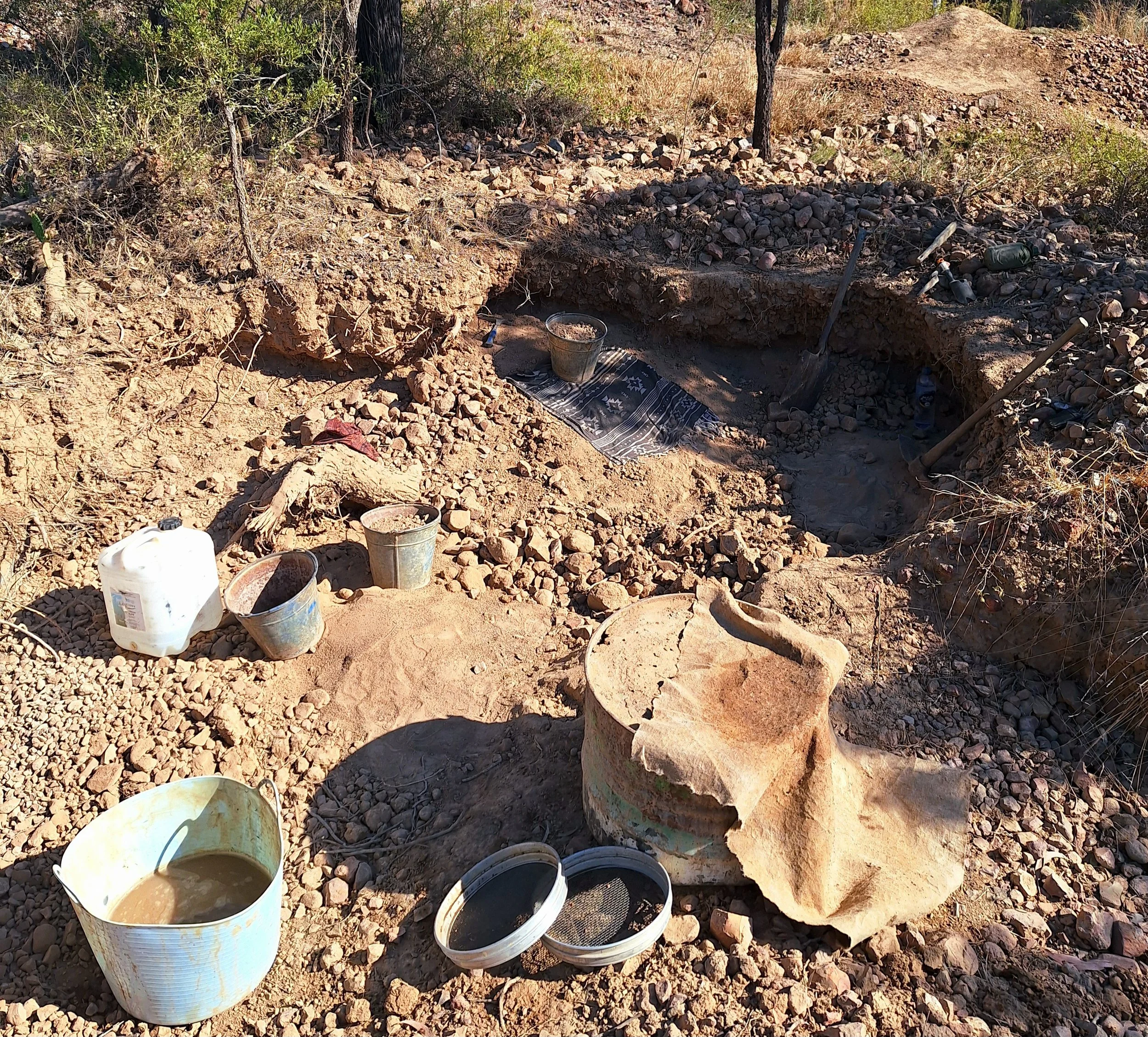 Archaeological excavation site with buckets, containers, shovels, and a large rock, surrounded by dry dirt and outdoor terrain.
