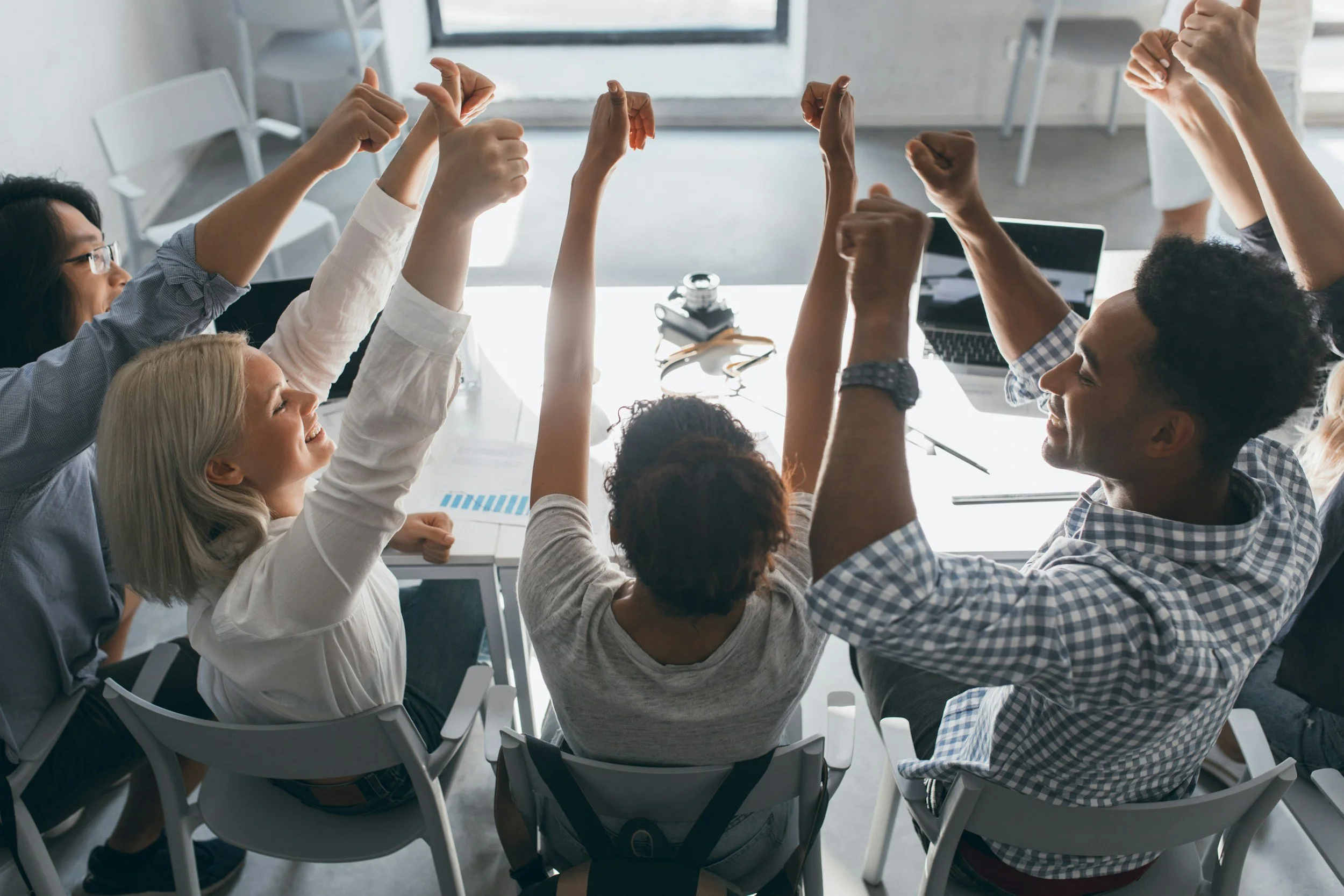 Group of diverse coworkers celebrating with arms raised in an office meeting room.