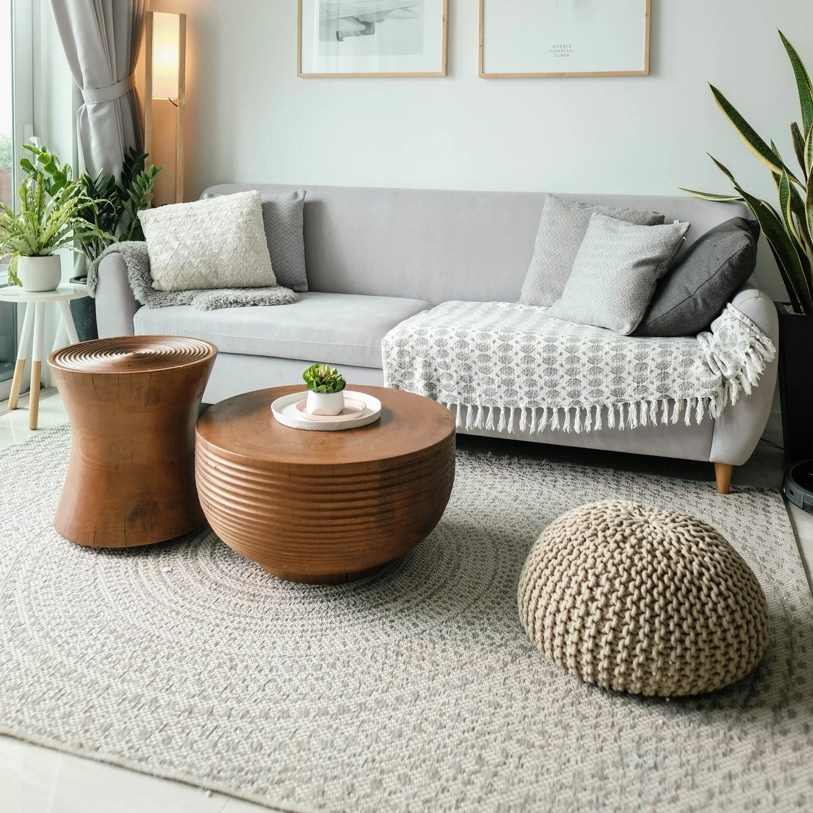 Living room with a light gray sofa adorned with various pillows, a knitted throw blanket, a beige pouf, wooden coffee tables, indoor plants, and wall art with a light-colored wall.