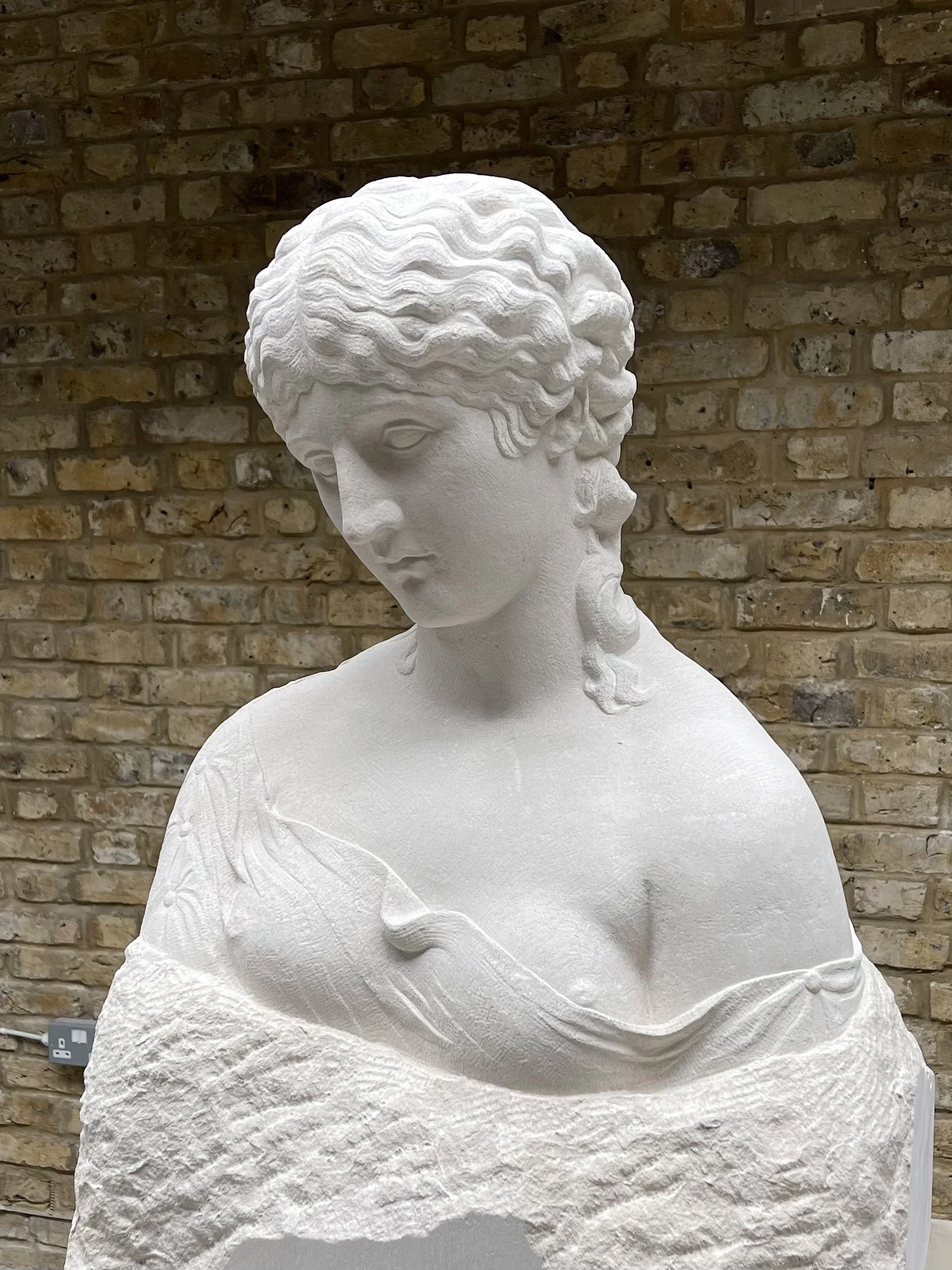 White marble statue of a woman with curly hair, wearing a draped garment, set against a brick wall.