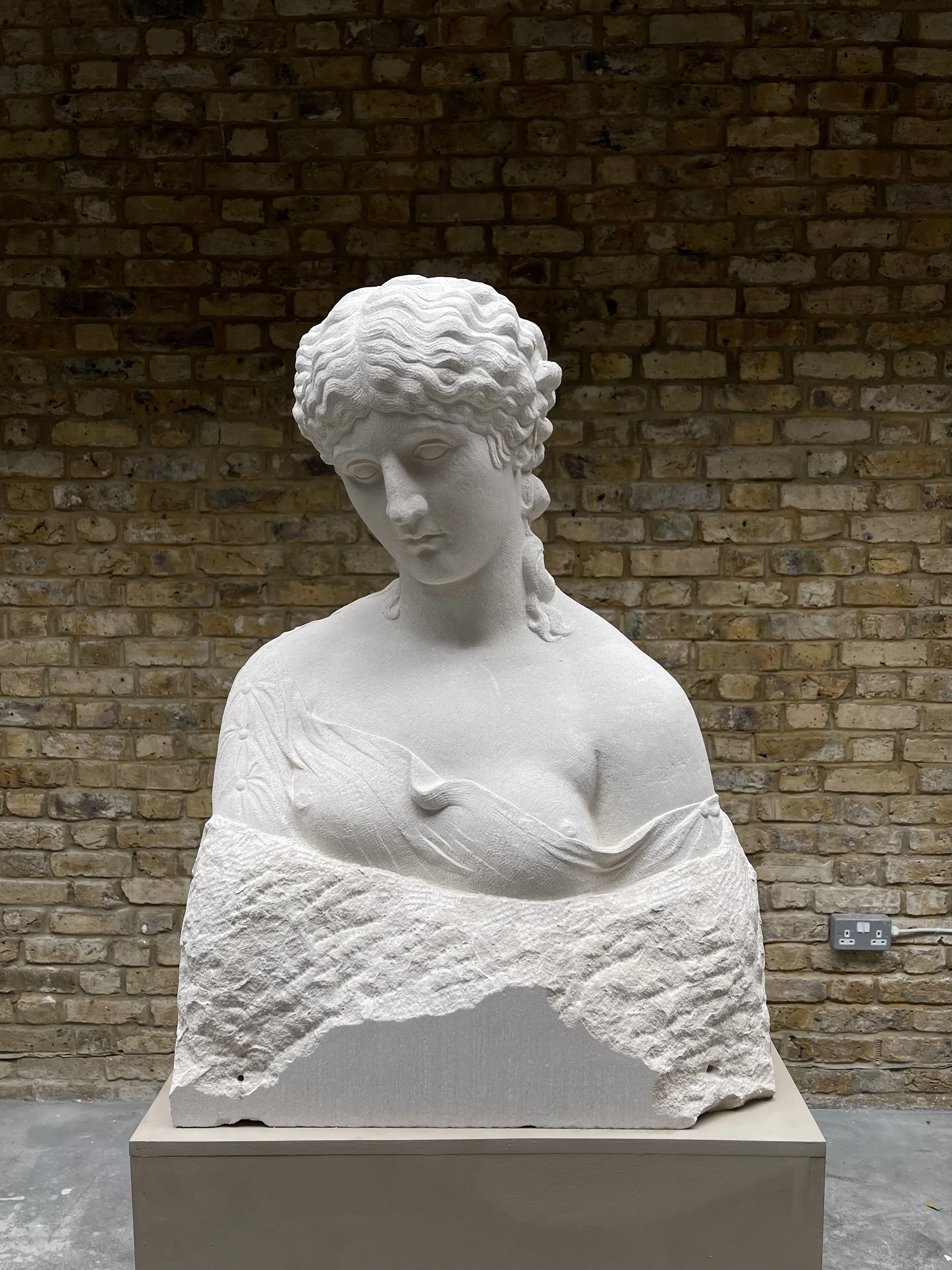 A white marble bust sculpture of a woman with curly hair, dressed in a draped garment, set against a brick wall background.