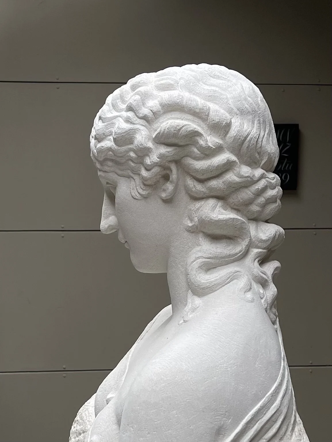 White sculpture of a woman with wavy hair, seen in profile, against a gray background.