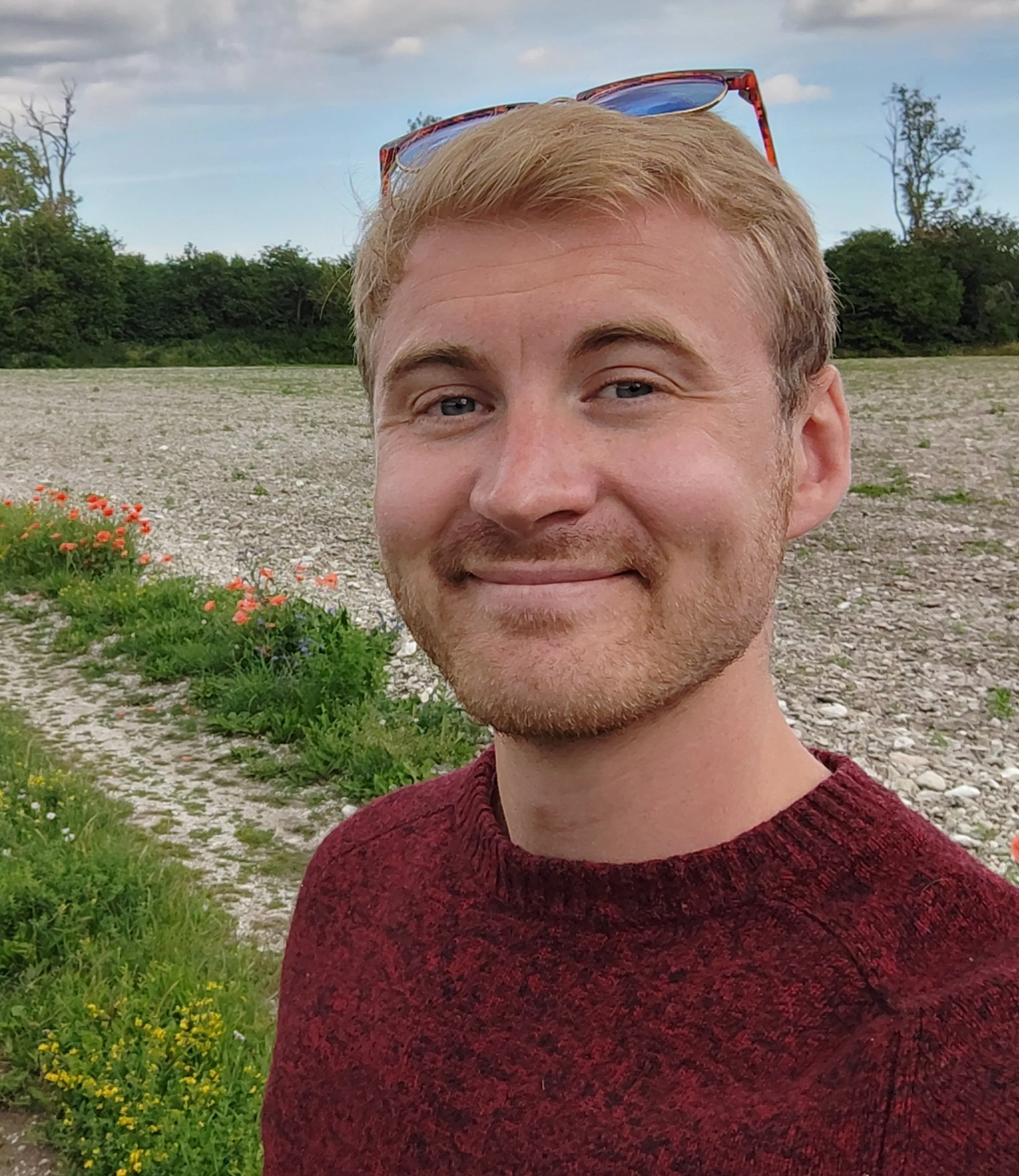 A young man with blonde hair, a beard, and sunglasses on his head, smiling outdoors in a natural setting with trees, plants, and a cloudy sky nearby.