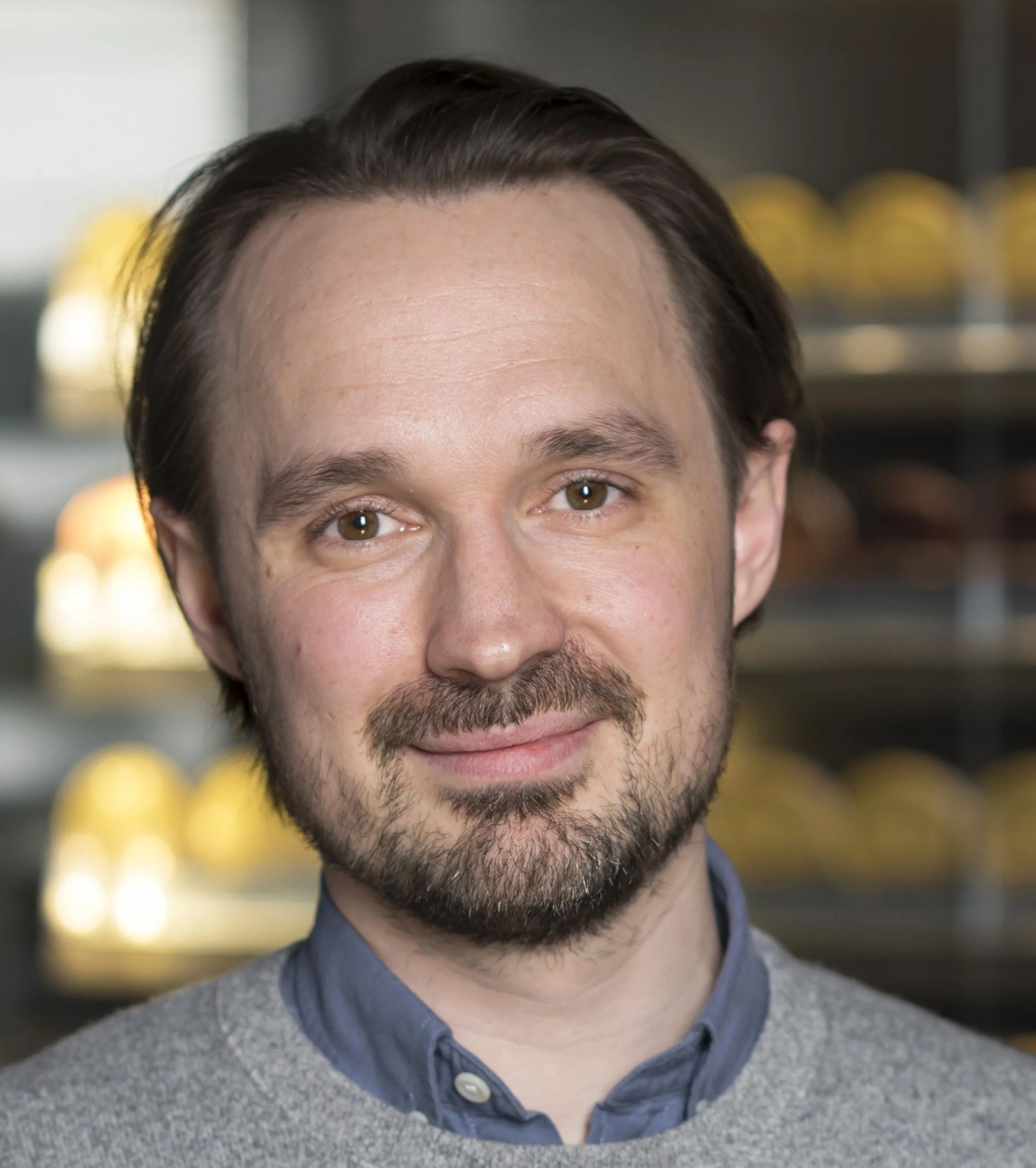 Close-up portrait of a man with brown hair, beard, and mustache, smiling in a setting with blurred yellow objects in the background.