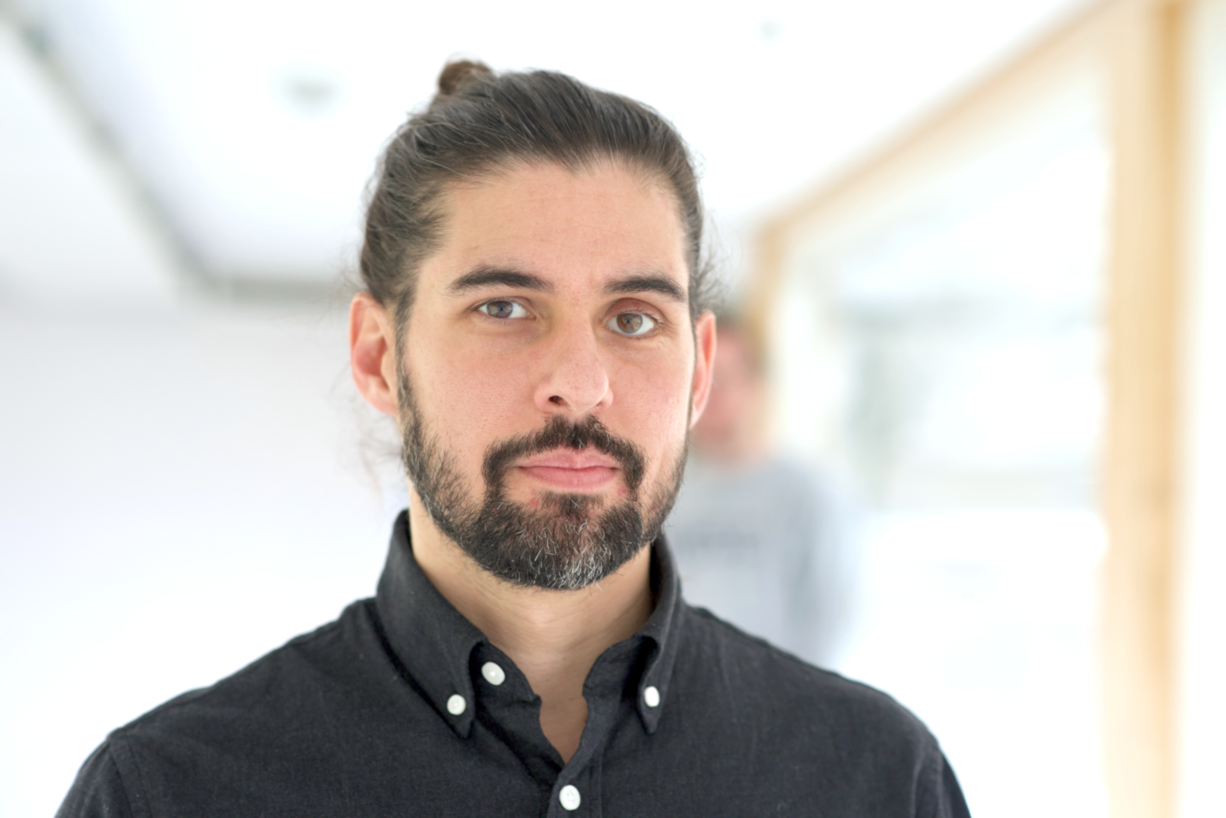 A man with dark hair, beard, and mustache, wearing a black button-up shirt, standing in a bright indoor environment with blurred background.