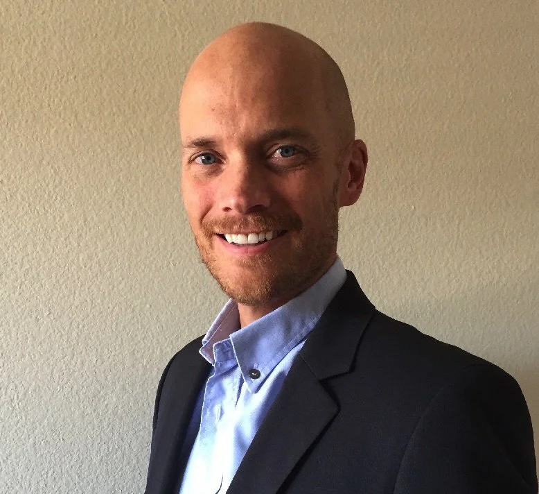 A bald man with a beard wearing a dark blazer and light blue dress shirt, smiling against a beige wall.