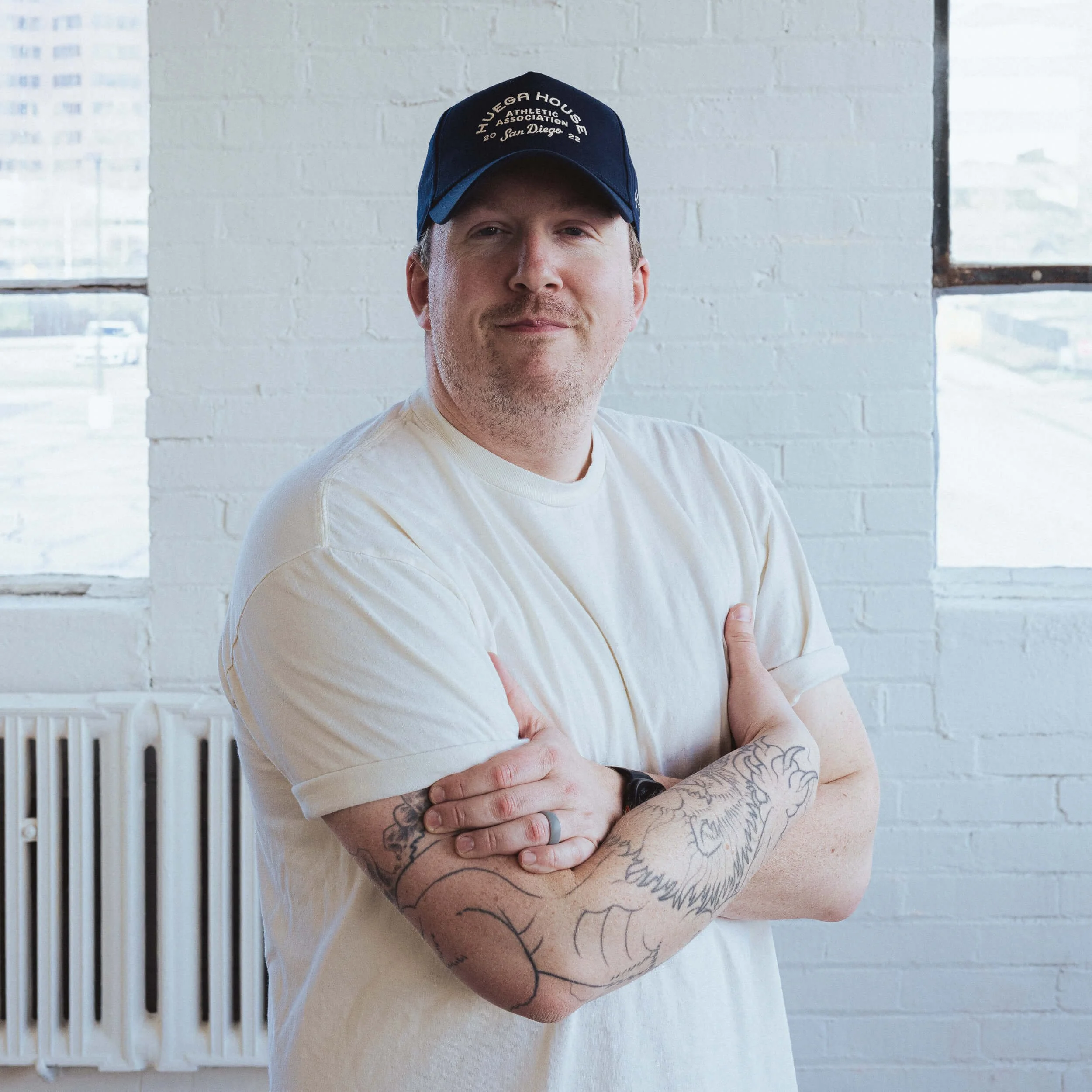 A young man with tattoos on his arms, wearing a beige t-shirt and a dark baseball cap, standing with crossed arms against a white brick wall with windows.