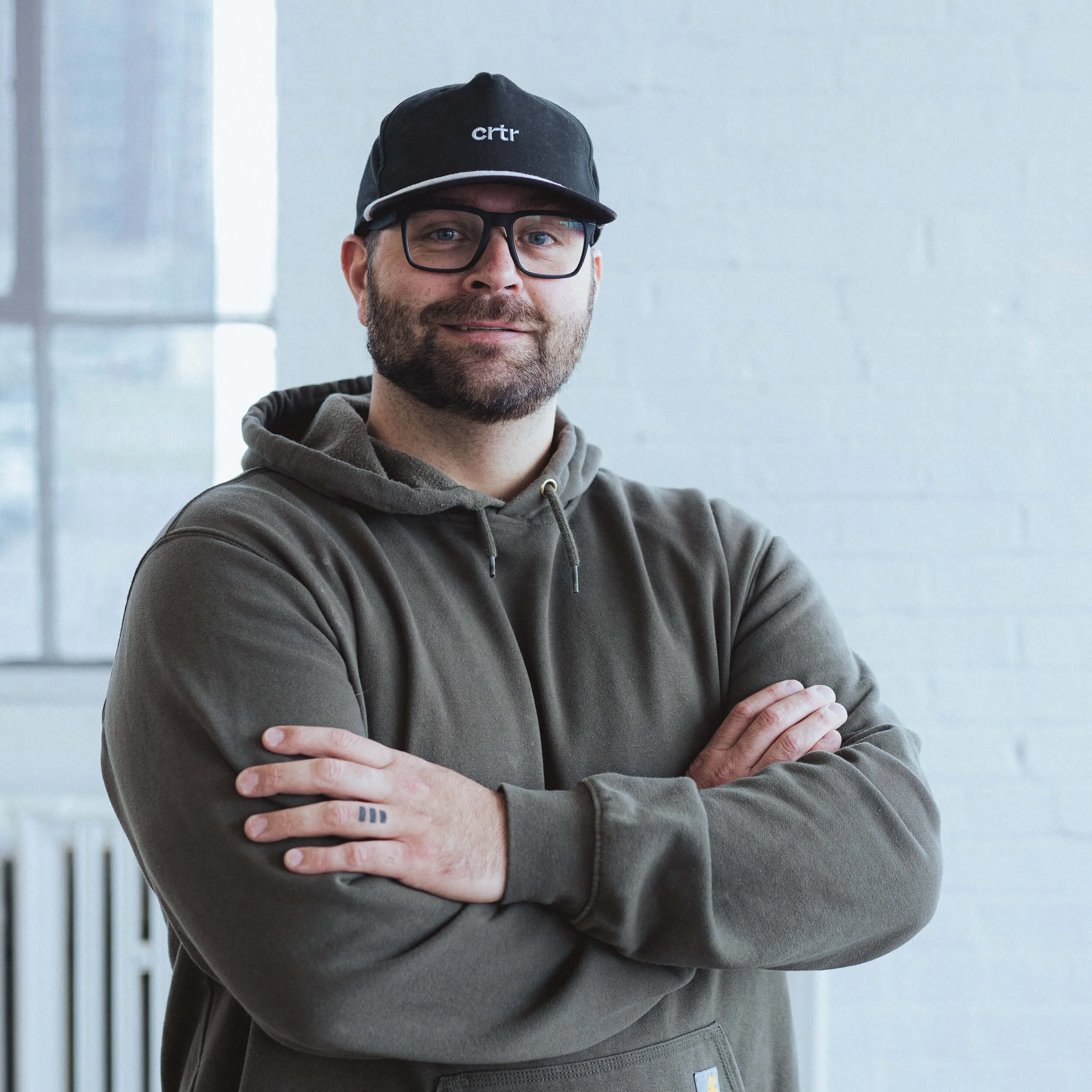 A man with glasses and a black baseball cap, wearing a hoodie, standing indoors with his arms crossed.